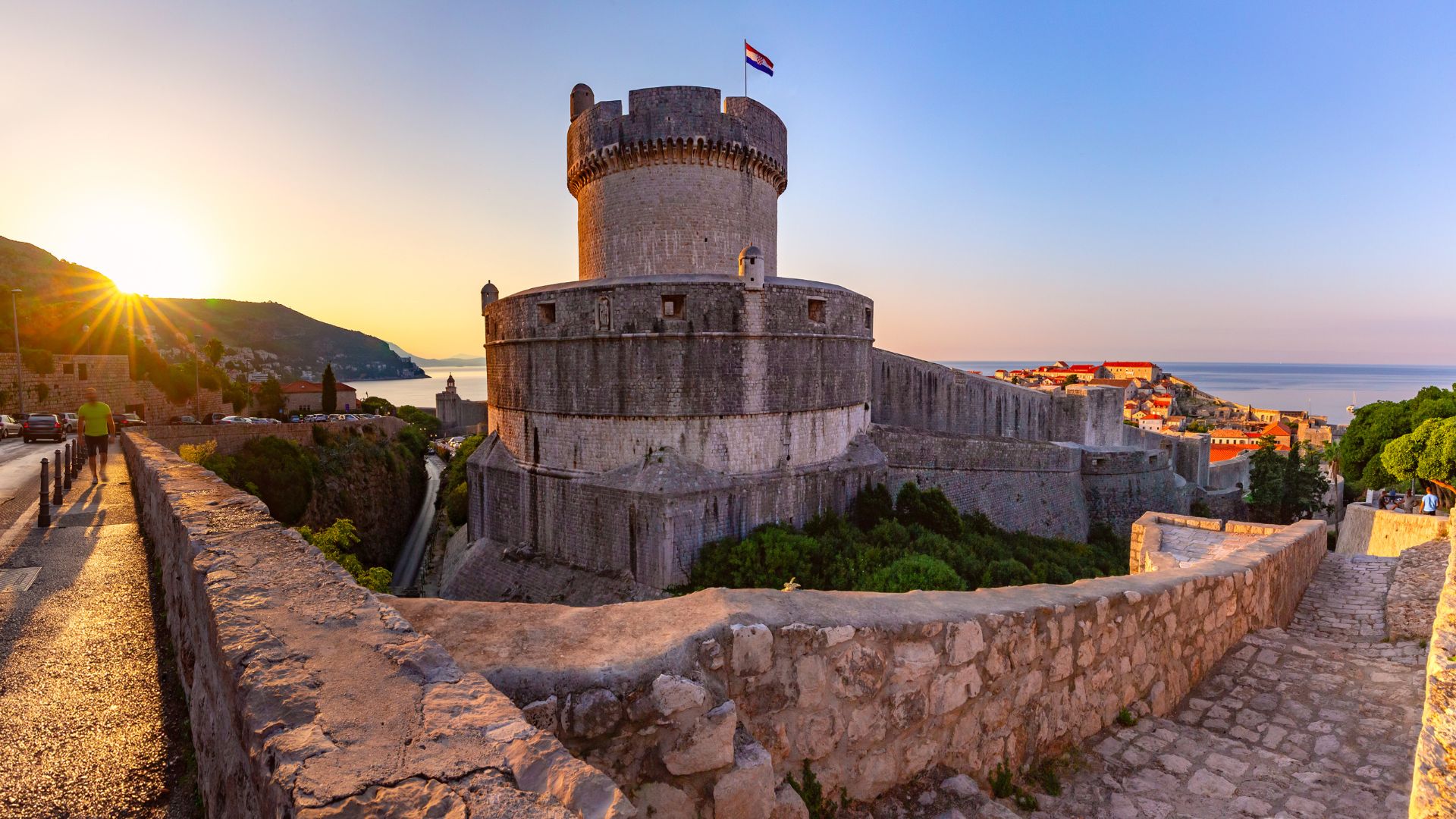 A stone fortress tower with a flag atop, part of ancient city walls, overlooking a city and the sea at sunset.