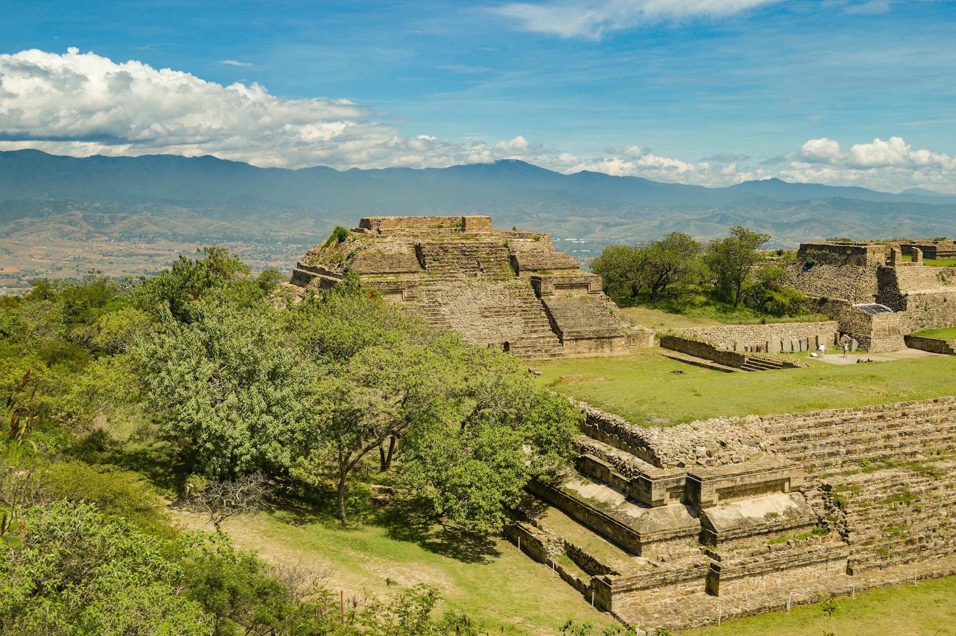 A panoramic view of Monte Albán, an ancient Zapotec city set atop a flattened mountaintop, with stone platforms, staircases, and pyramids spread across a wide ceremonial plaza