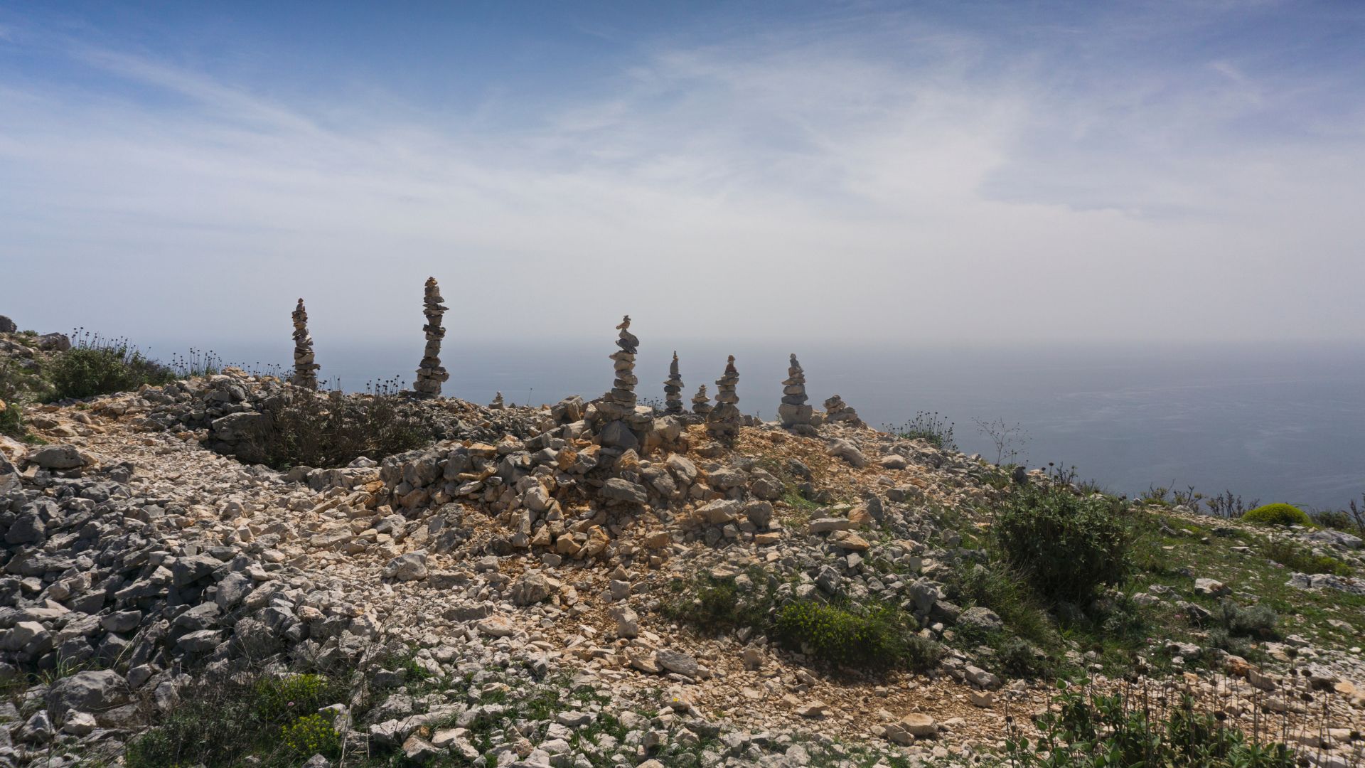 A wide-angle view of a rocky, arid hilltop covered in loose stones and sparse vegetation, with several prominent cairns (stacked stone pillars) standing against a hazy sky and the distant, blurred expanse of the sea.