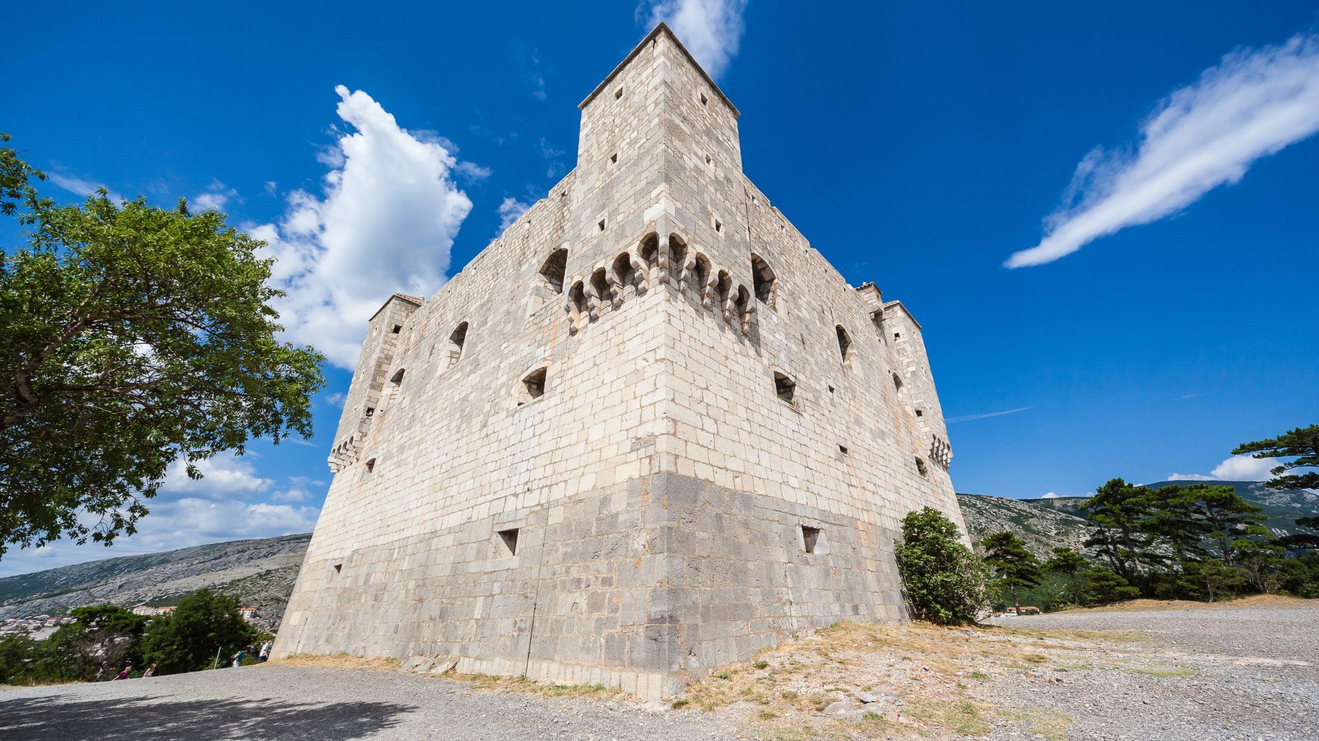 A low-angle view captures the imposing, square-shaped Nehaj Fortress in Senj, Croatia, under a bright blue sky with scattered white clouds. The fortress, made of light-colored stone, stands prominently on a hill with a few trees visible around its base and mountains in the background. 