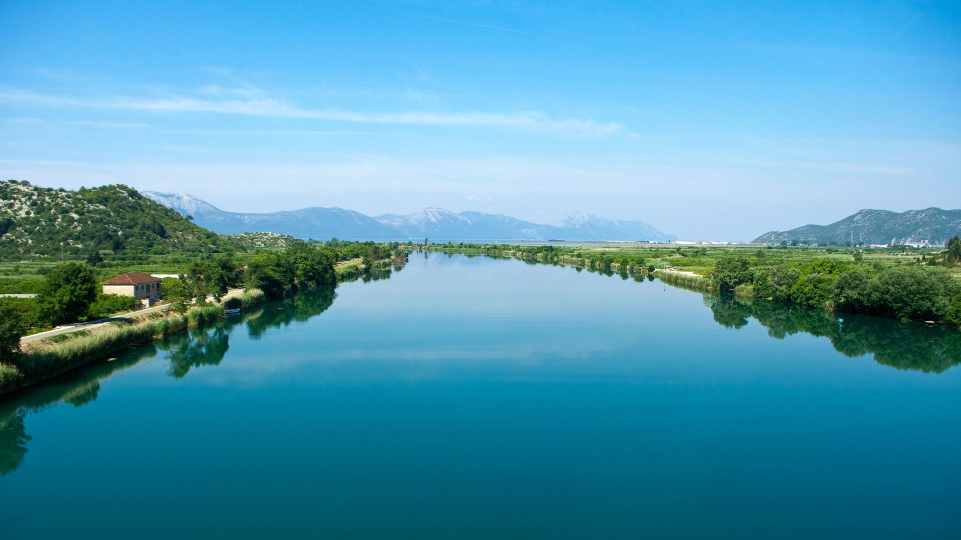 A wide, calm river with turquoise water flows between lush green banks, lined with trees and distant mountains under a clear blue sky. On the left bank, a small building is visible amidst the greenery.