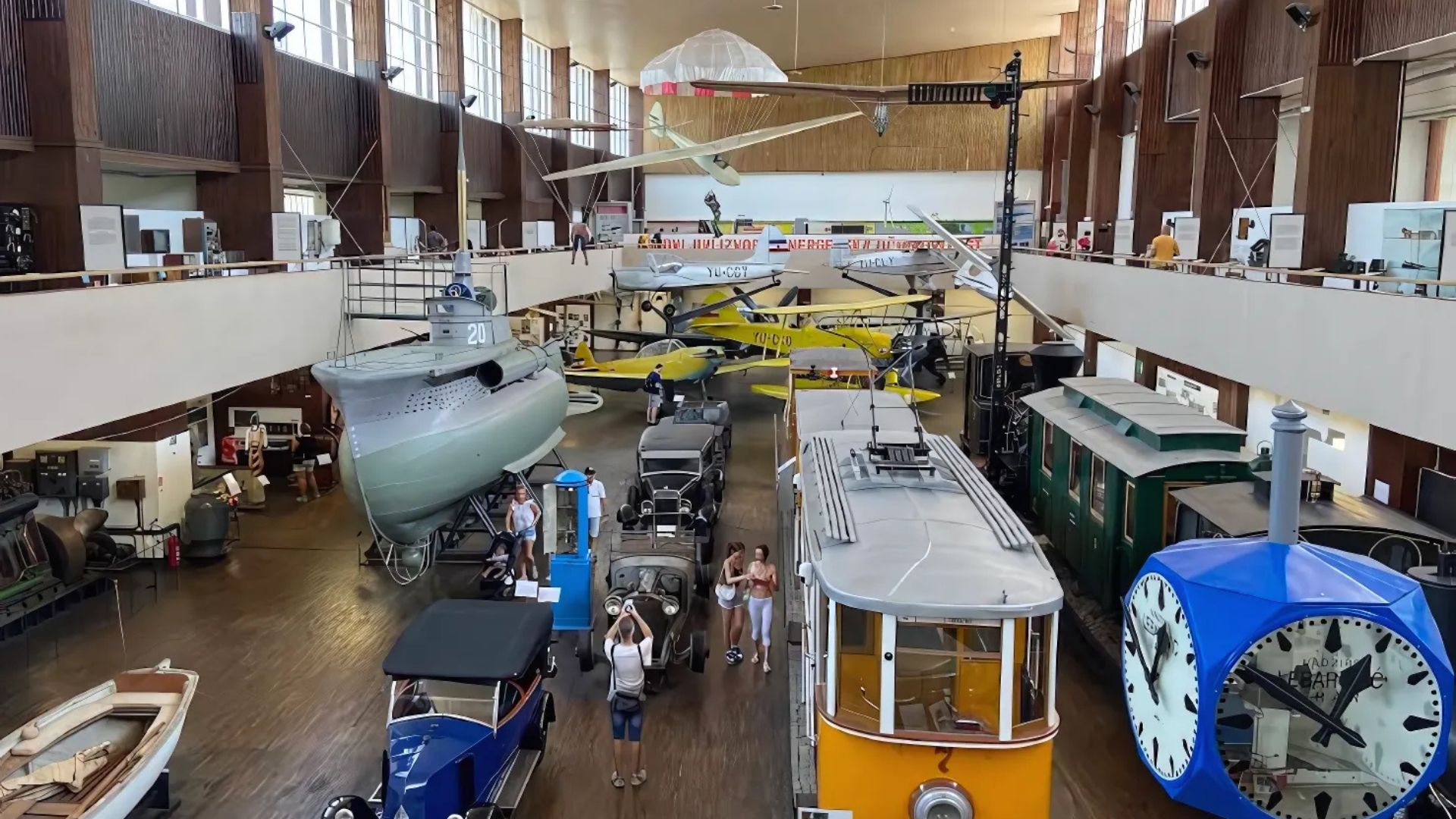 An indoor shot of a large museum hall displaying various historical technological exhibits, including a submarine, multiple vintage cars, a tram, and several aircraft suspended from the ceiling, with visitors observing the displays.