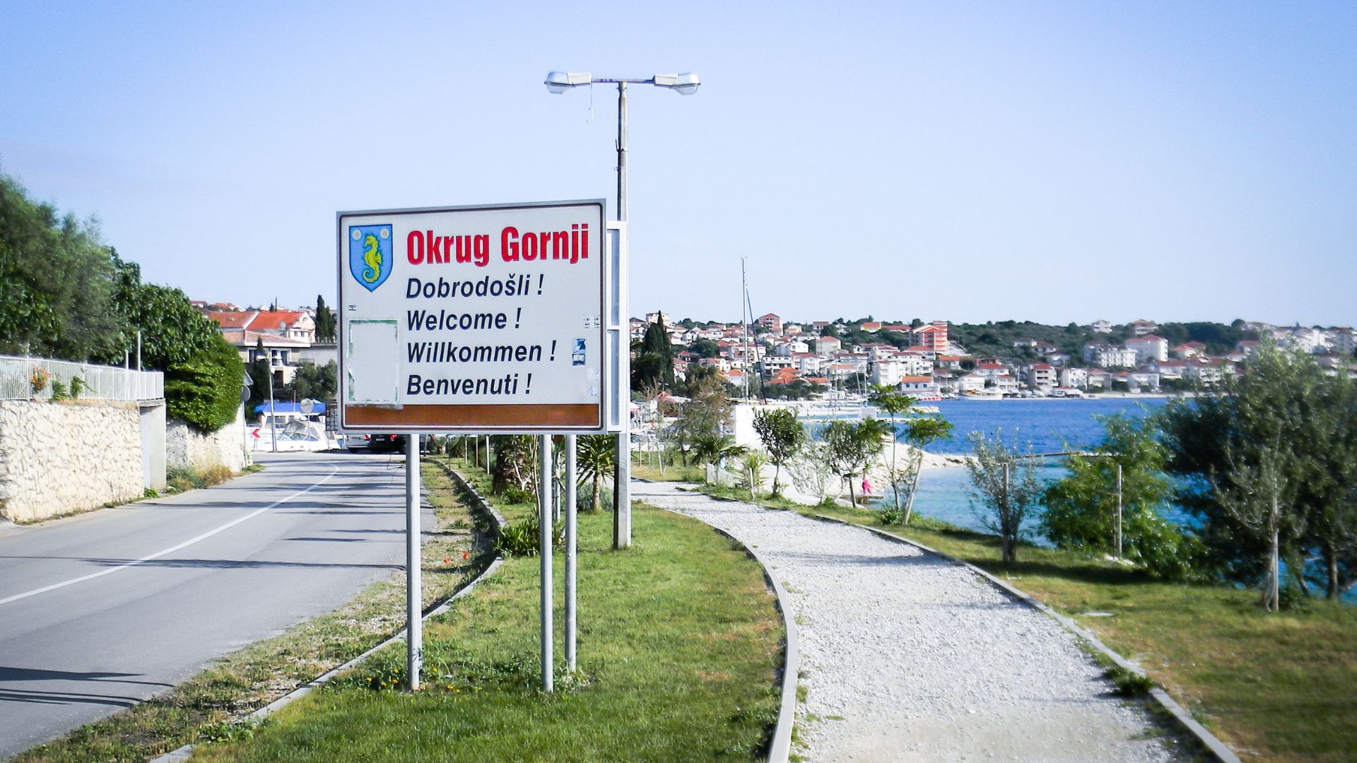 A roadside sign for "Okrug Gornji" in multiple languages (Croatian, English, German, Italian) stands beside a paved path and a road, with a coastal town and the sea visible in the sunny background.