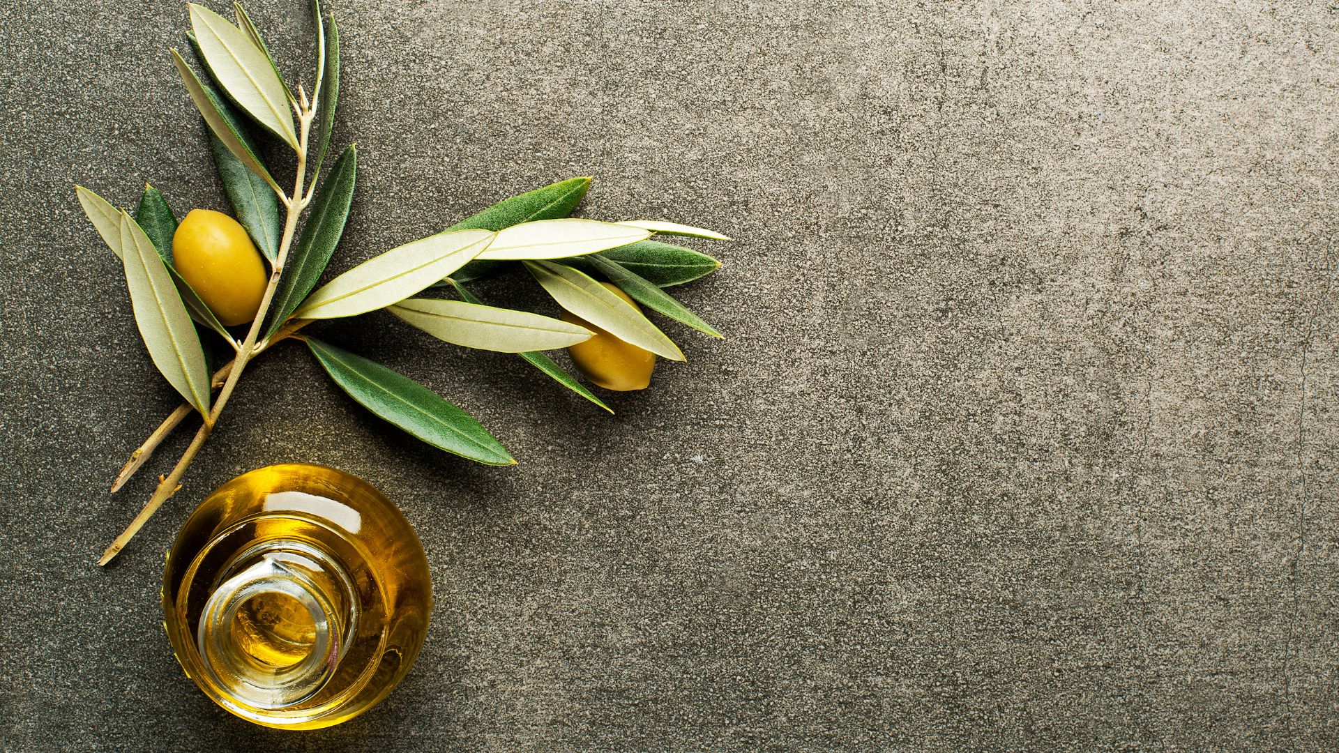 A close-up, overhead shot of an olive branch with green leaves and two ripe, yellowish-green olives, alongside a small, clear glass bottle filled with golden olive oil, all resting on a dark gray, textured surface. 