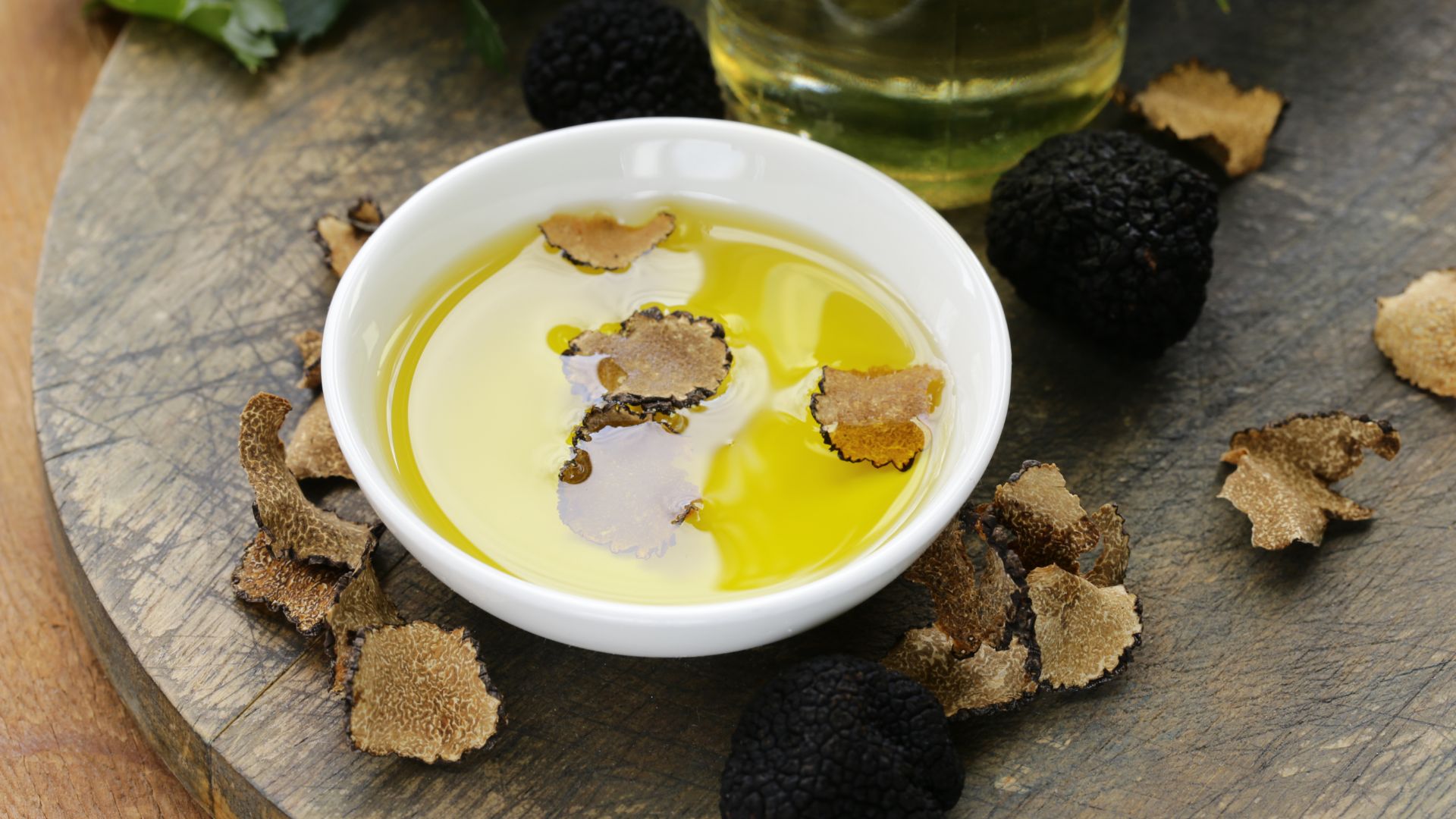 A white bowl of golden truffle oil with truffle slices, surrounded by whole black truffles and more truffle pieces on a rustic wooden surface.