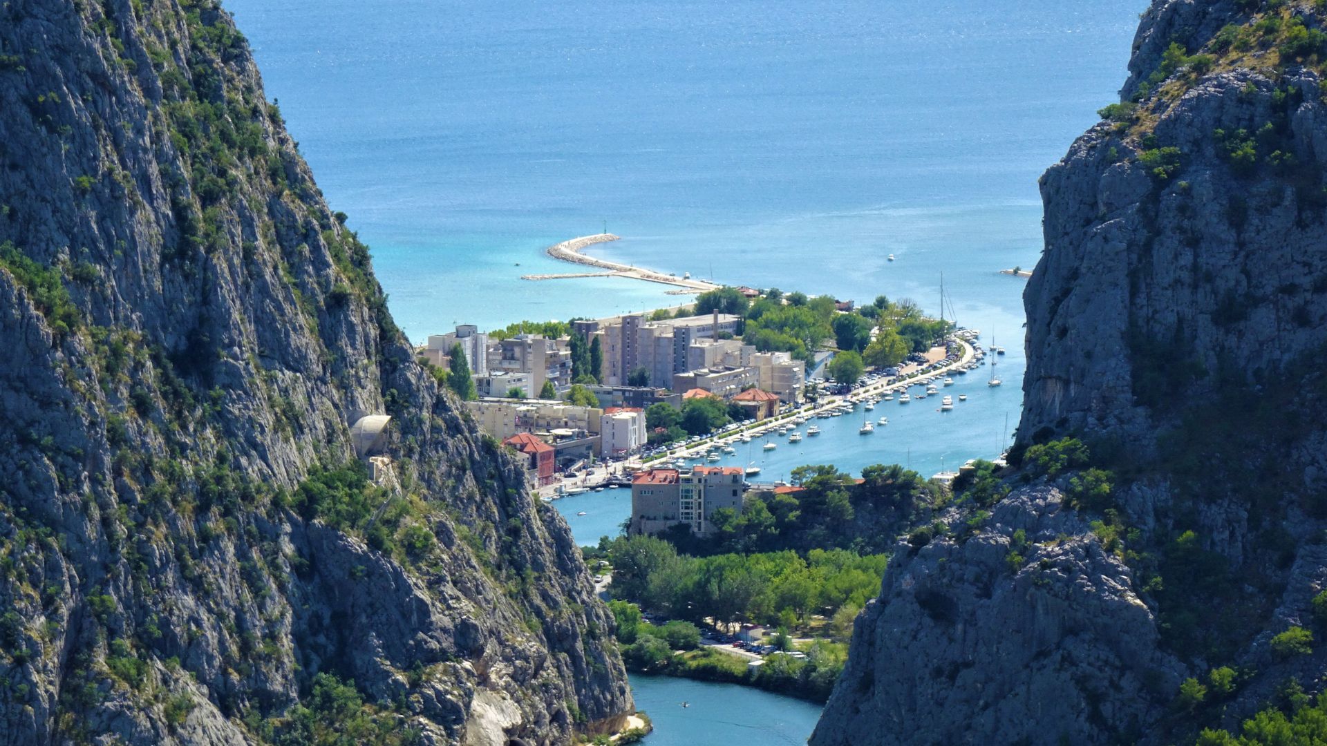 A panoramic view of Omiš, Croatia, nestled between towering rocky cliffs, with the Cetina River flowing into the Adriatic Sea, where boats are docked along the waterfront and a town with buildings is visible along the coastline and on a small peninsula.