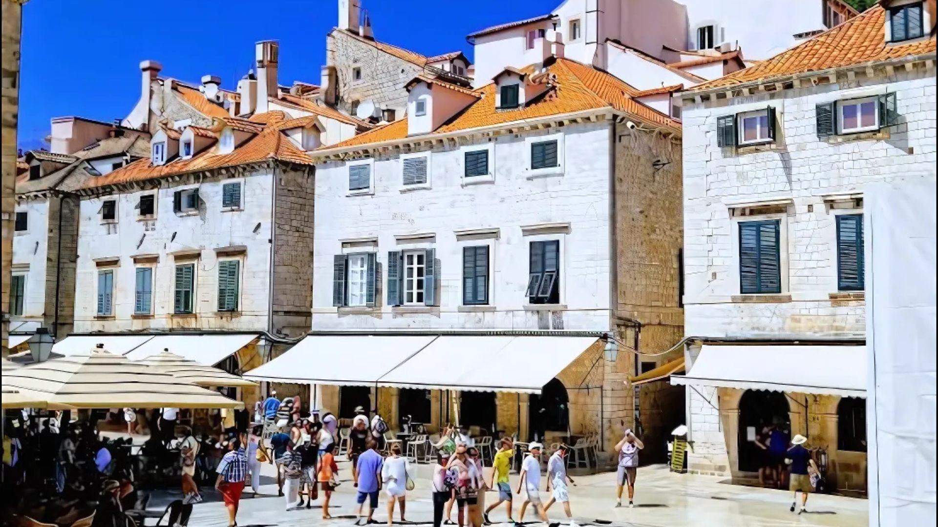 A daytime view of Luža Square in Dubrovnik, Croatia, showing a bustling crowd of people gathered in the sunlit square surrounded by historic stone buildings with red-tiled roofs and green-shuttered windows, along with awnings extending over ground-level establishments. 