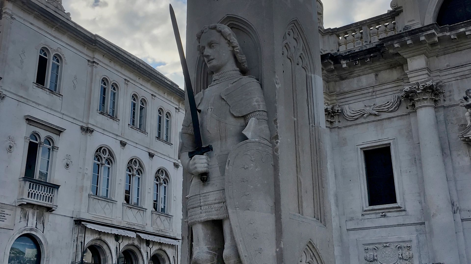 A stone statue of a knight in armor, known as Orlando's Column, stands prominently in a public square surrounded by historic European buildings with arched windows and balconies under a cloudy sky.