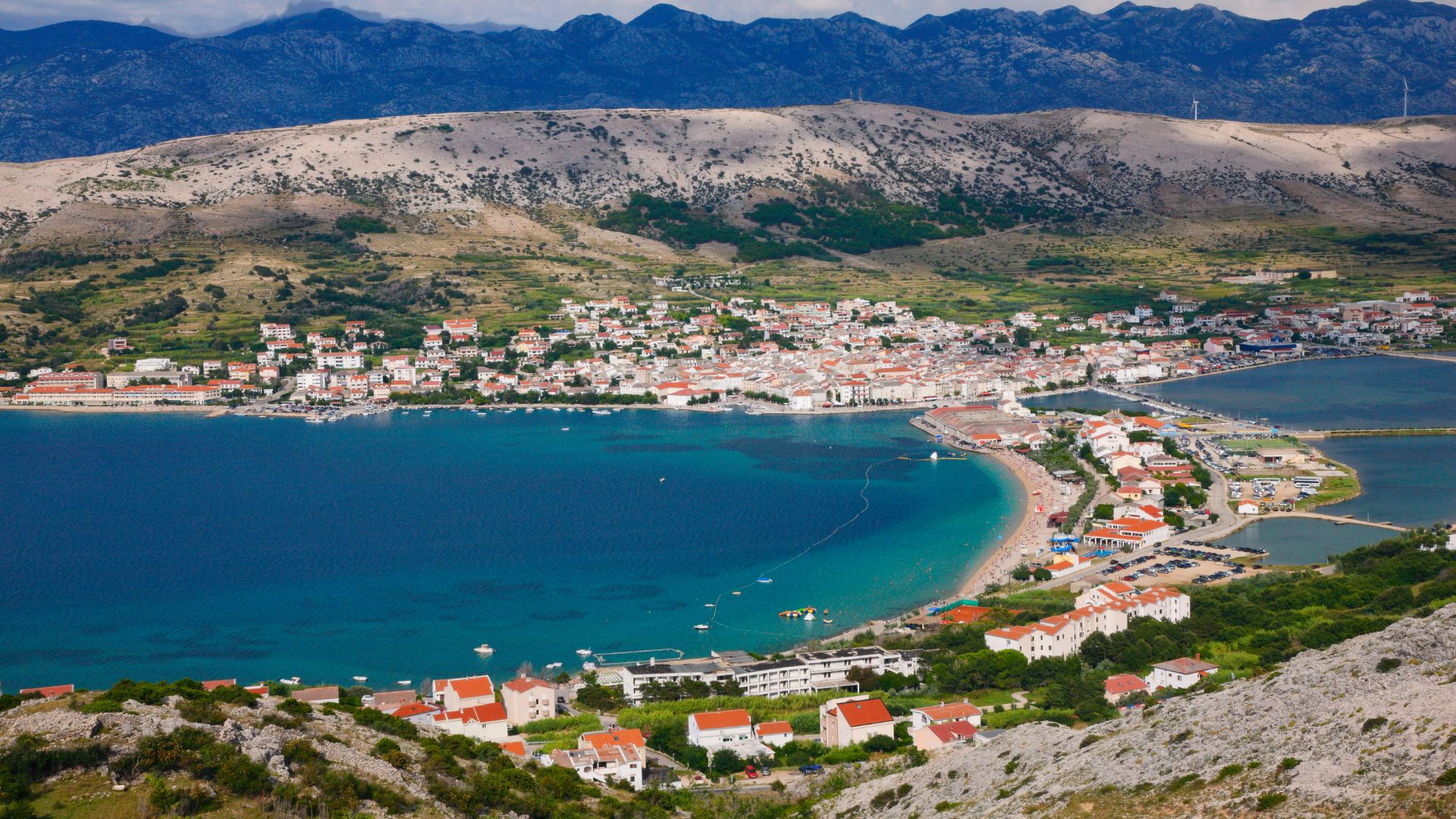 A panoramic view of the town of Pag on Pag Island, Croatia, showing a vibrant blue bay with boats, a sandy beach, and a densely built coastal town nestled against a backdrop of barren, rocky hills under a partly cloudy sky.