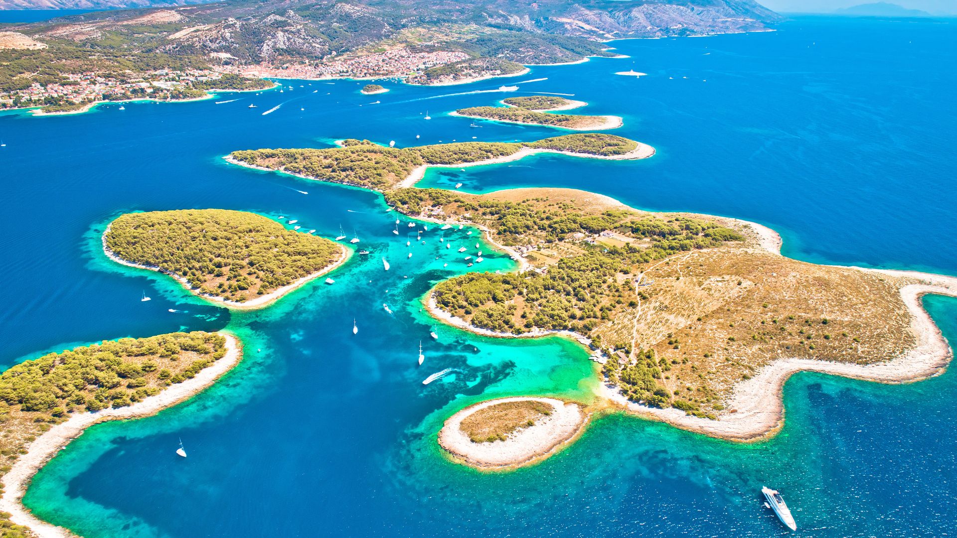 An aerial view showcases the Pakleni Islands, a cluster of lush, green islands and islets surrounded by clear turquoise and deep blue waters of the Adriatic Sea, with numerous boats dotting the bays and channels.