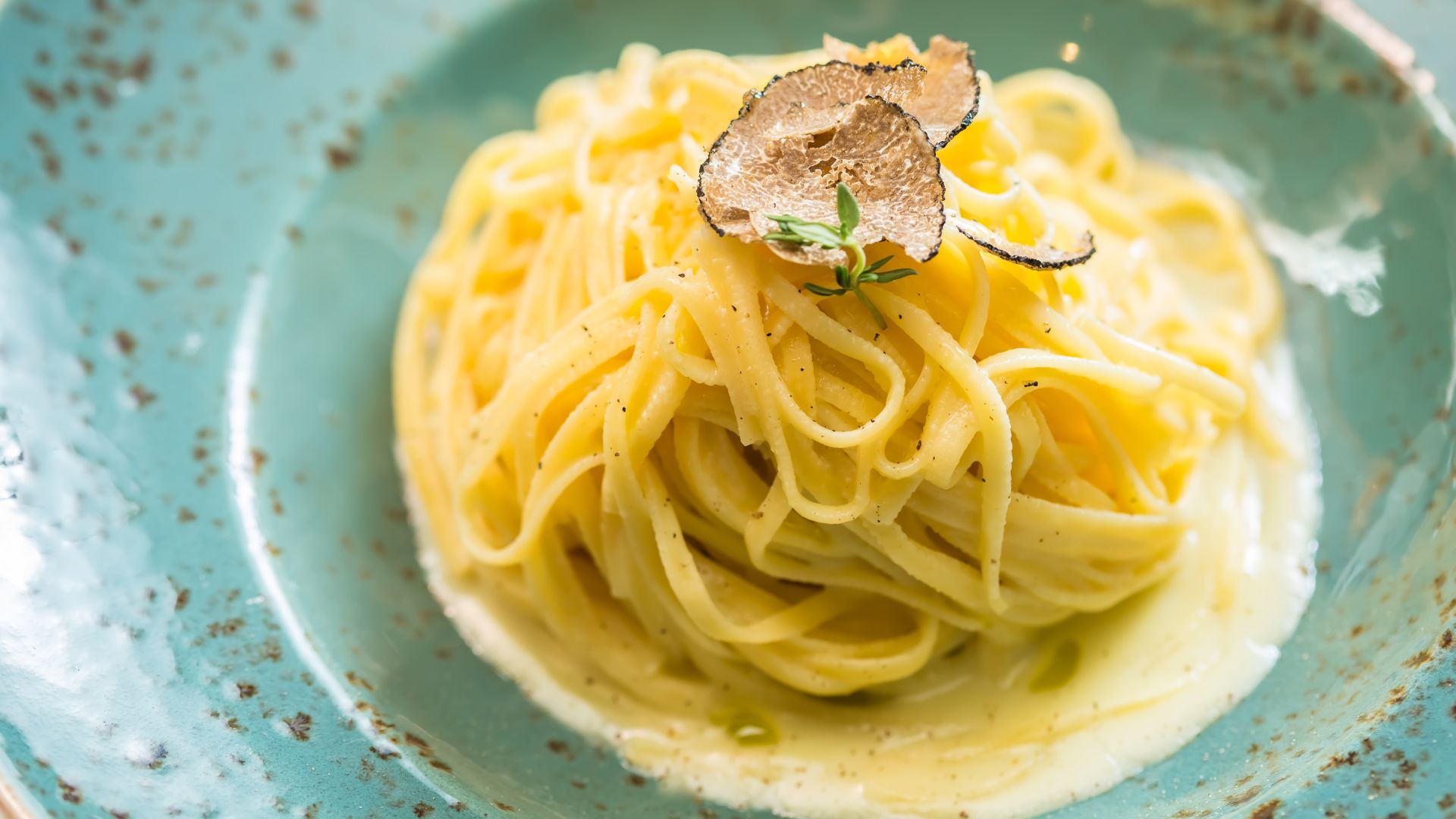 A close-up of a plate of pasta, likely tagliolini or spaghetti, topped with delicate shavings of white truffle and a creamy sauce.