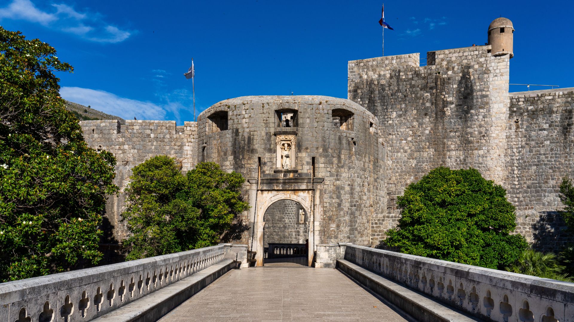 A stone bridge leads to the imposing Pile Gate, the main entrance to Dubrovnik's historic walled city, under a clear blue sky.