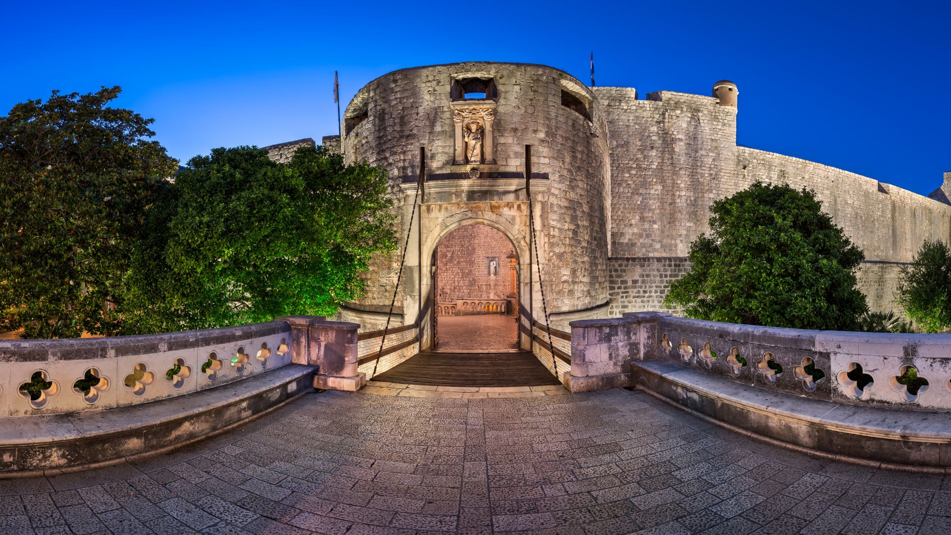 A panoramic shot of the historic Pile Gate in Dubrovnik, Croatia, at dusk, featuring its stone archway, drawbridge, and surrounding walls, flanked by lush green trees under a deep blue sky.
