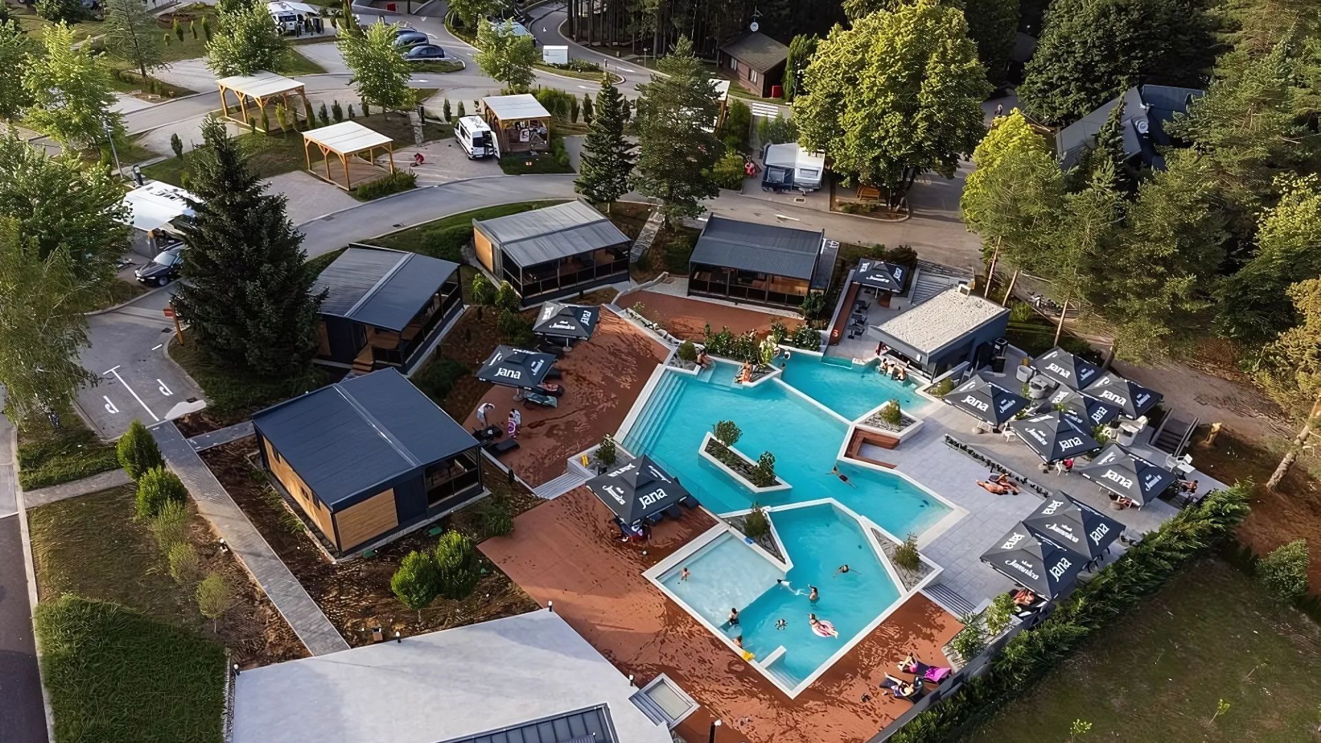 An aerial view of the Plitvice Holiday Resort, showcasing a complex of modern cabins and mobile homes nestled among trees, surrounding a multi-level outdoor swimming pool area with sun loungers and umbrellas. Campervans and cars are visible in parking areas nearby.