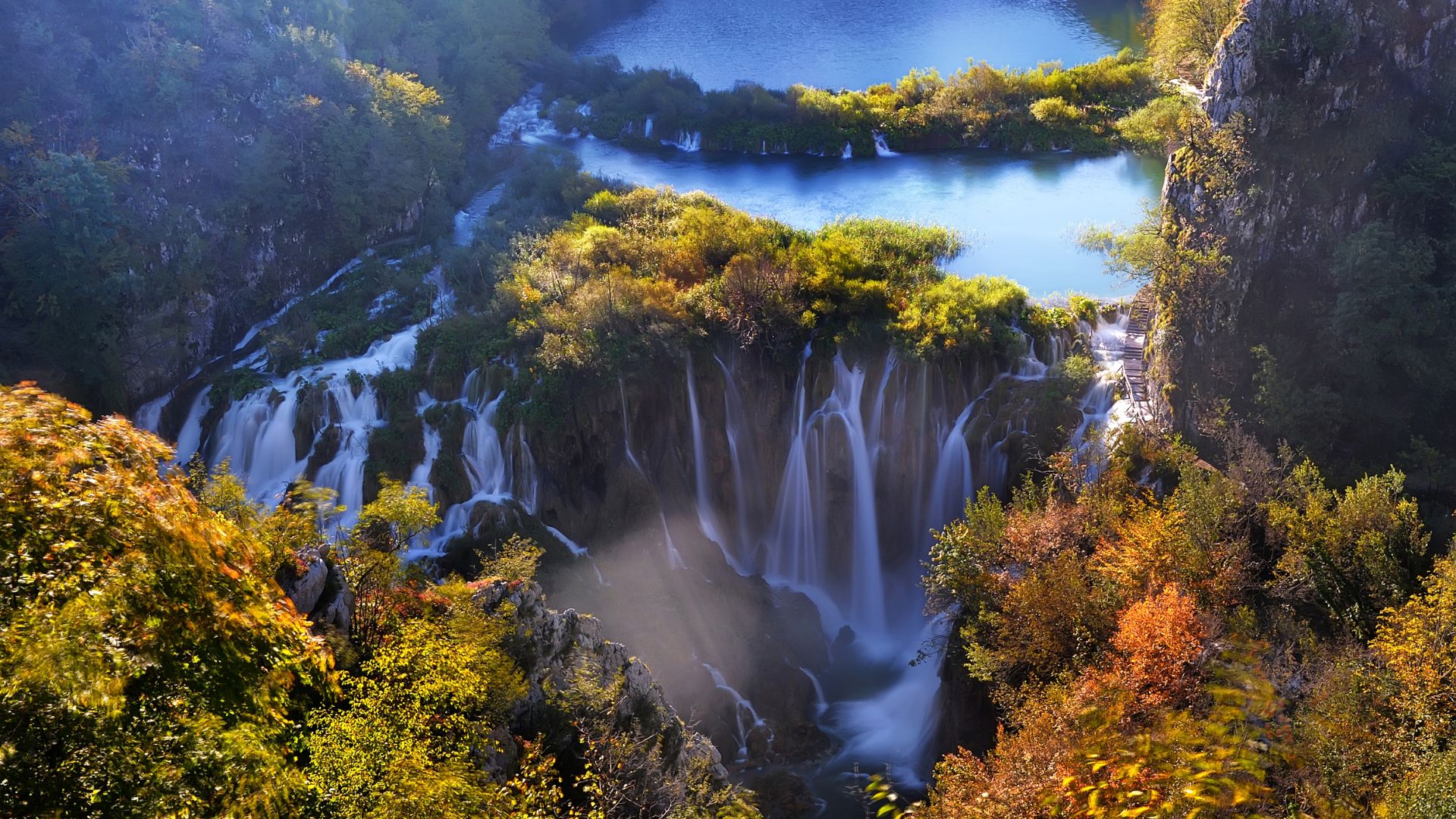 A panoramic view of the cascading waterfalls and turquoise lakes of Plitvice Lakes National Park, surrounded by lush, colorful autumn foliage under a bright sky.