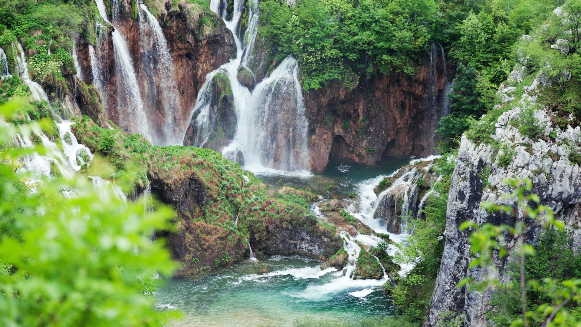  A lush, green landscape features multiple cascading waterfalls flowing into turquoise lakes, surrounded by dense foliage and rocky cliffs within Plitvice Lakes National Park.
