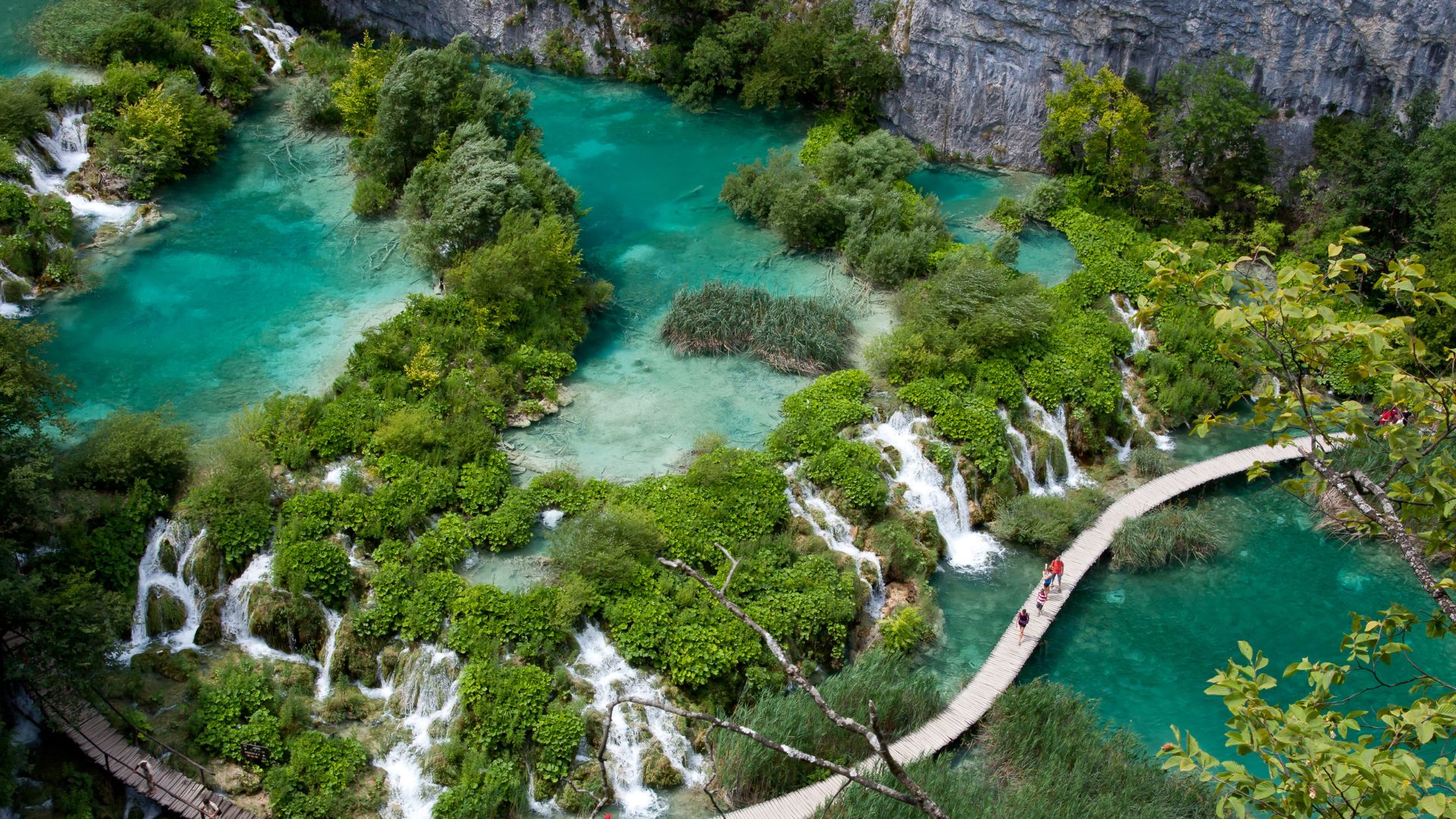 An aerial view captures the vibrant turquoise waters and cascading waterfalls of Plitvice Lakes National Park, surrounded by lush green foliage and a winding wooden boardwalk with visitors strolling along it.