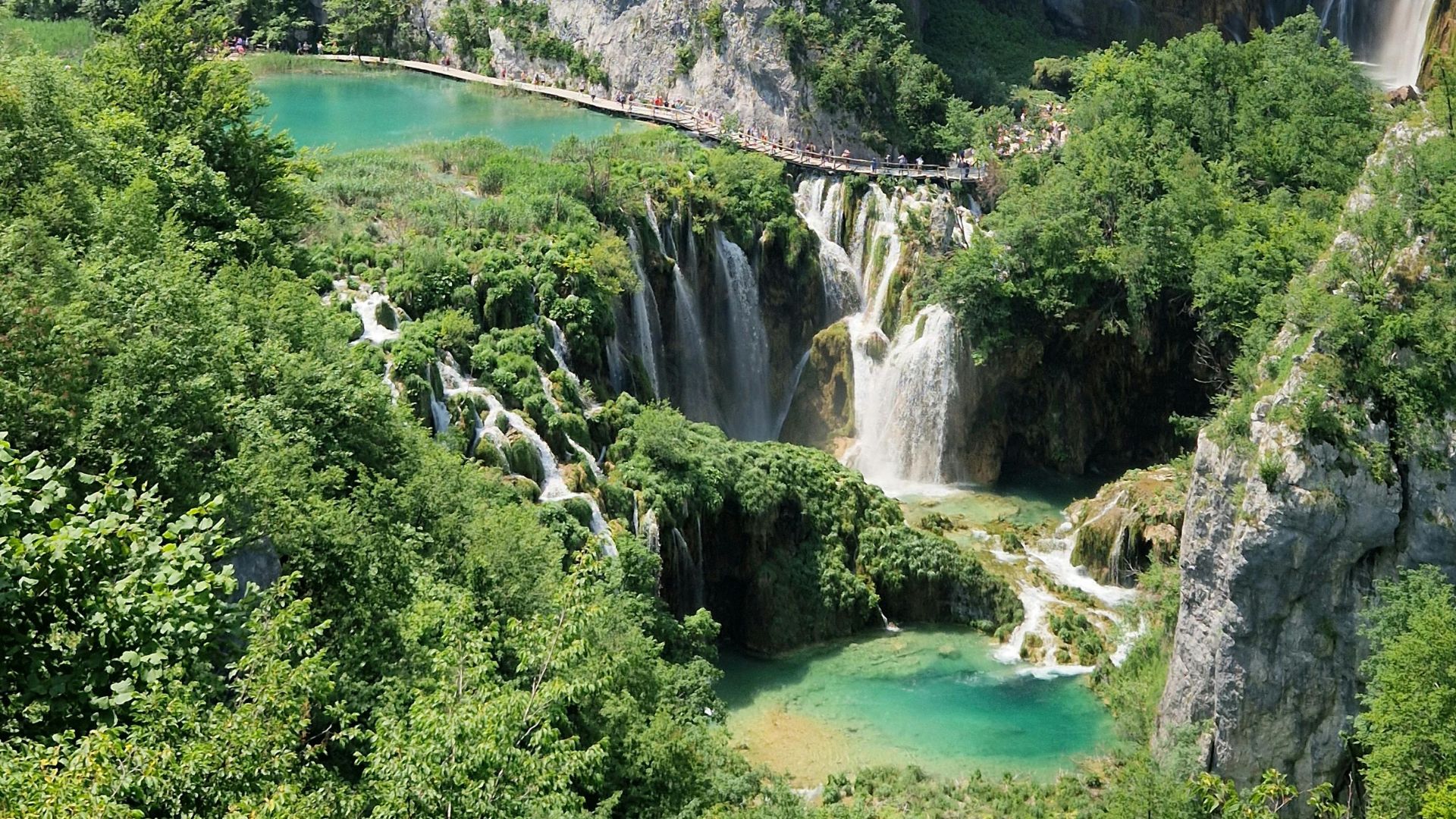 An aerial view of Plitvice Lakes National Park in Croatia, showing vibrant turquoise lakes and multiple waterfalls cascading through lush green forests and rock formations, with a wooden walkway visible winding along the upper lake and waterfalls. 