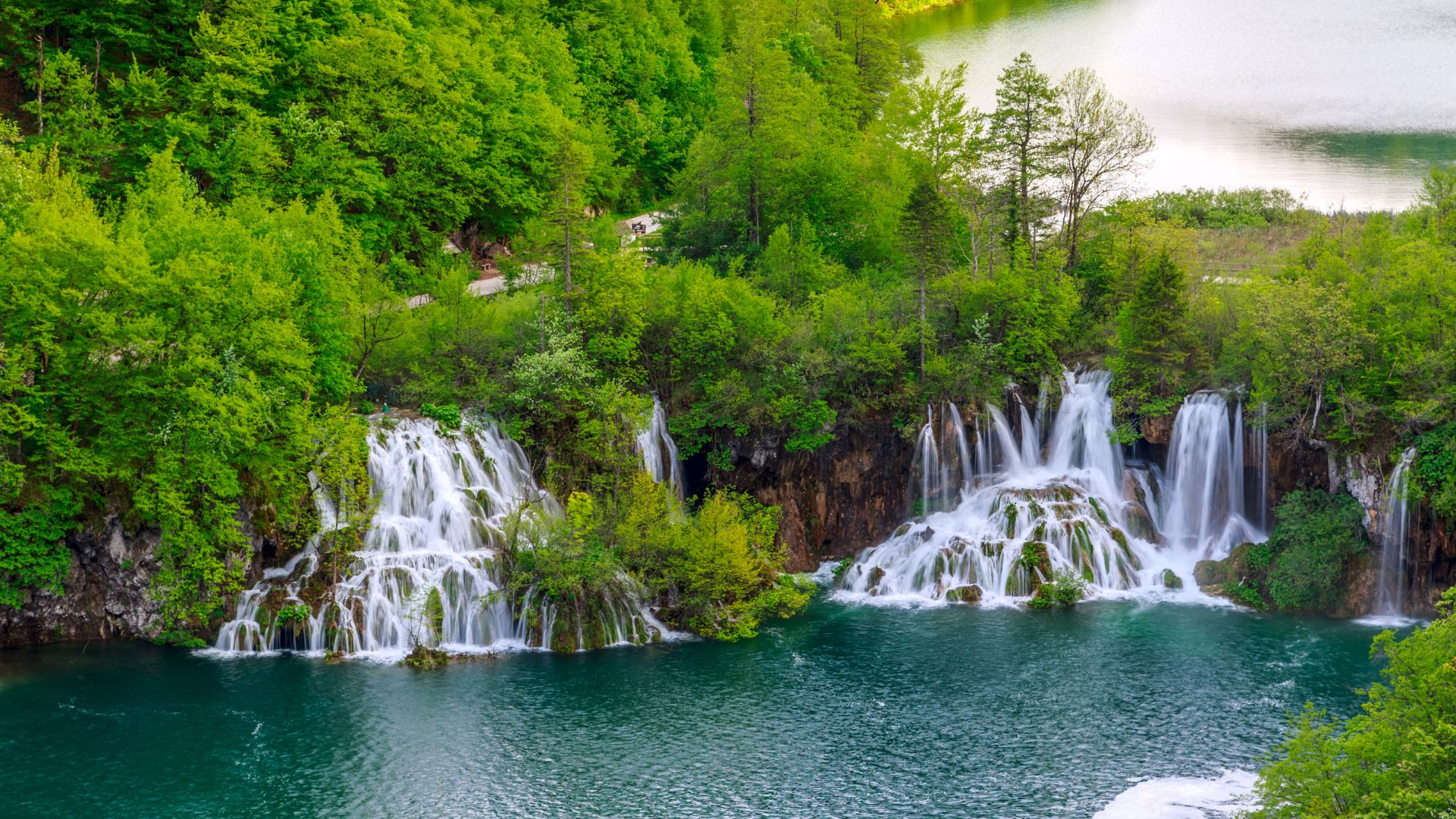 A vibrant landscape featuring multiple cascading waterfalls flowing into a clear, turquoise lake, surrounded by lush green forests under a bright sky.