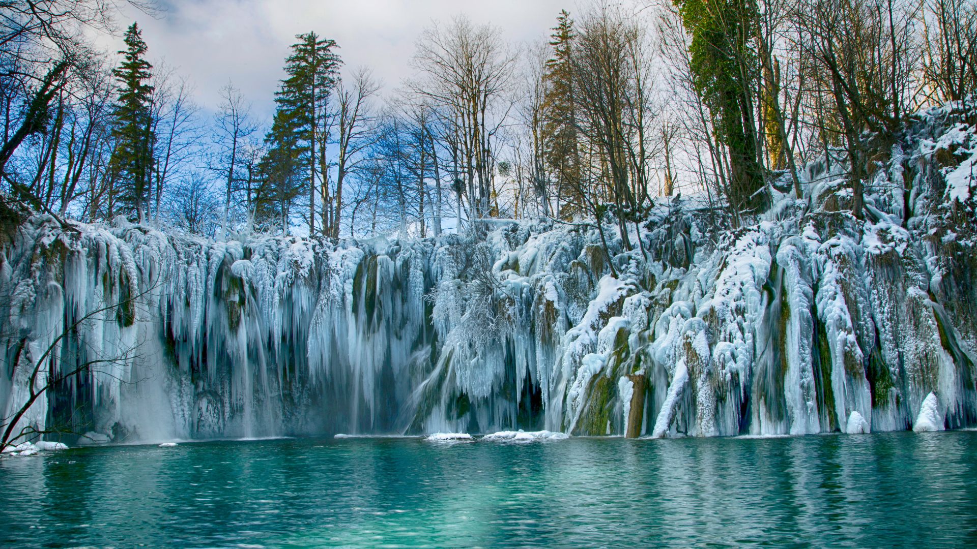 A winter scene at Plitvice Lakes National Park showing a series of frozen waterfalls cascading into a vibrant turquoise lake, surrounded by snow-covered trees and bare branches.