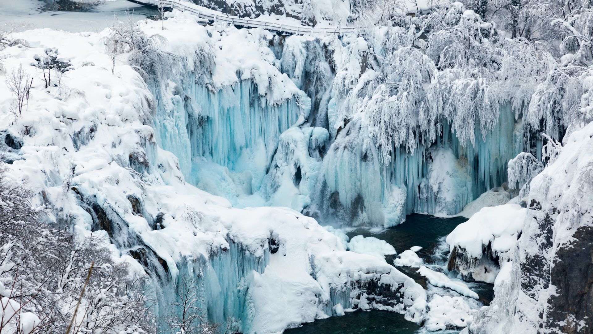 A stunning winter scene at Plitvice Lakes National Park, featuring snow-covered landscapes, frozen waterfalls with prominent blue-tinted icicles, and partially frozen turquoise lakes. 