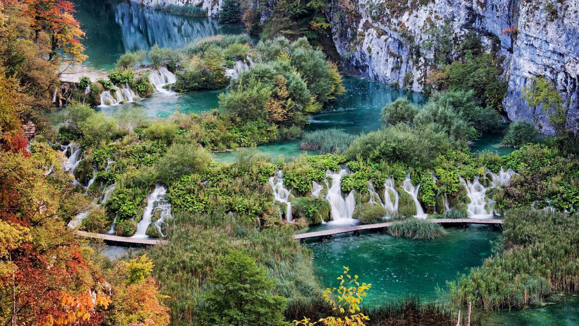 A panoramic view of Plitvice Lakes National Park, showcasing a series of interconnected turquoise lakes and cascading waterfalls amidst lush green and autumnal foliage, with wooden walkways winding through the water.