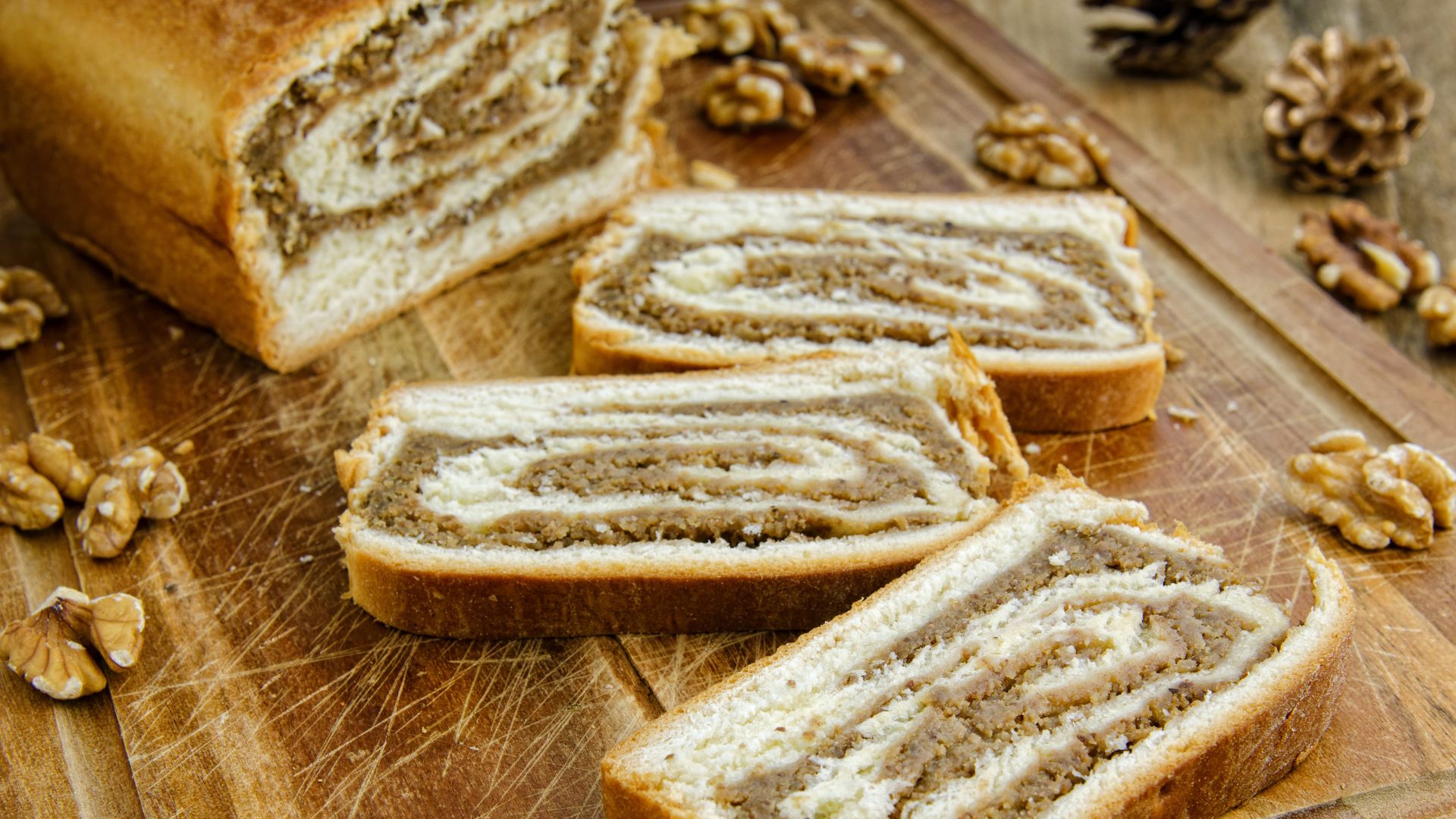 A close-up of a sliced nut roll on a wooden cutting board, revealing its distinctive spiral pattern of dough and walnut filling, with whole walnuts scattered around the roll and slices. 
