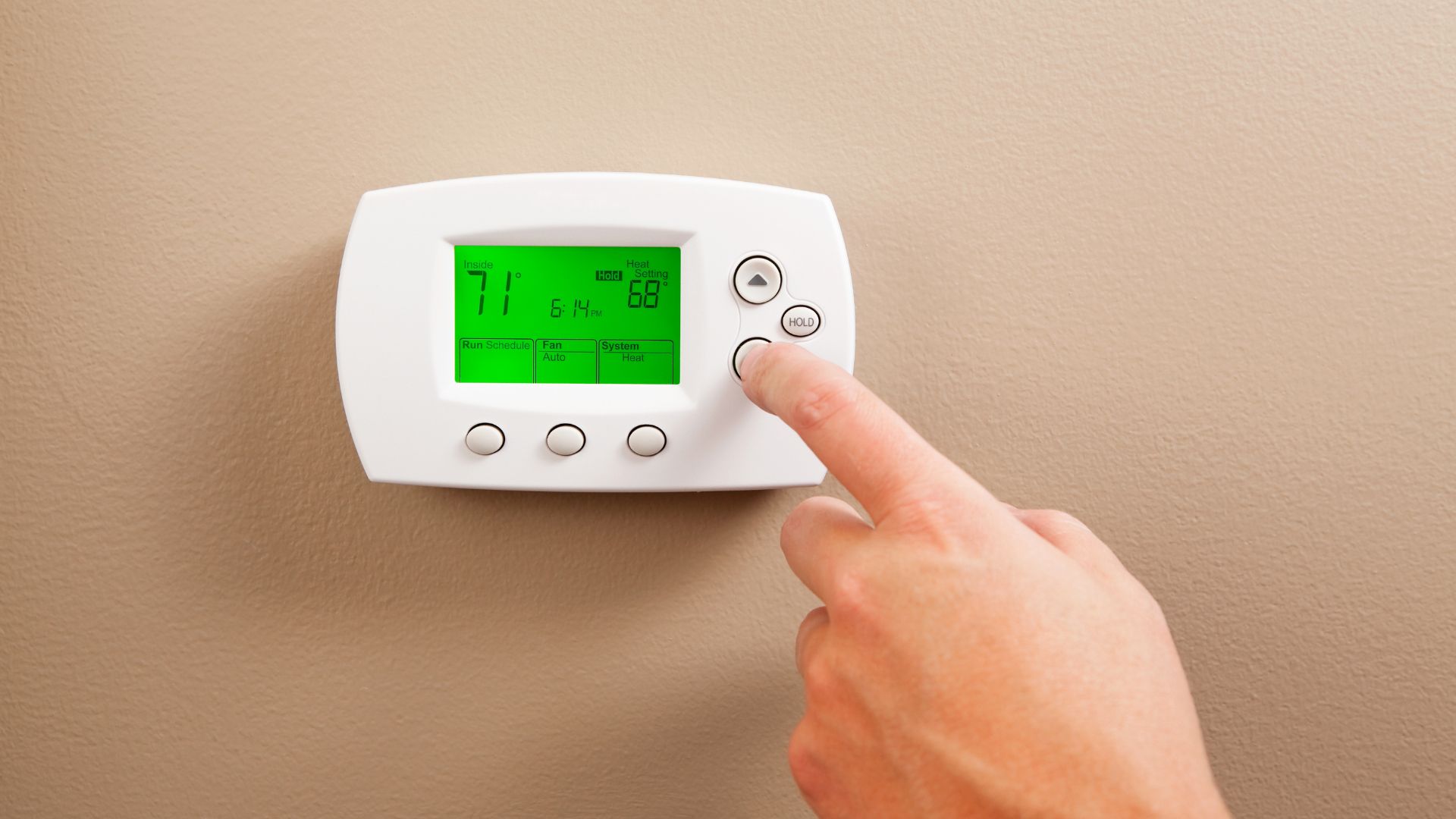 A close-up shows a white digital thermostat mounted on a beige wall, with a green backlit display showing "71" and "68". A person's finger is pressing one of the control buttons on the right side of the device.
