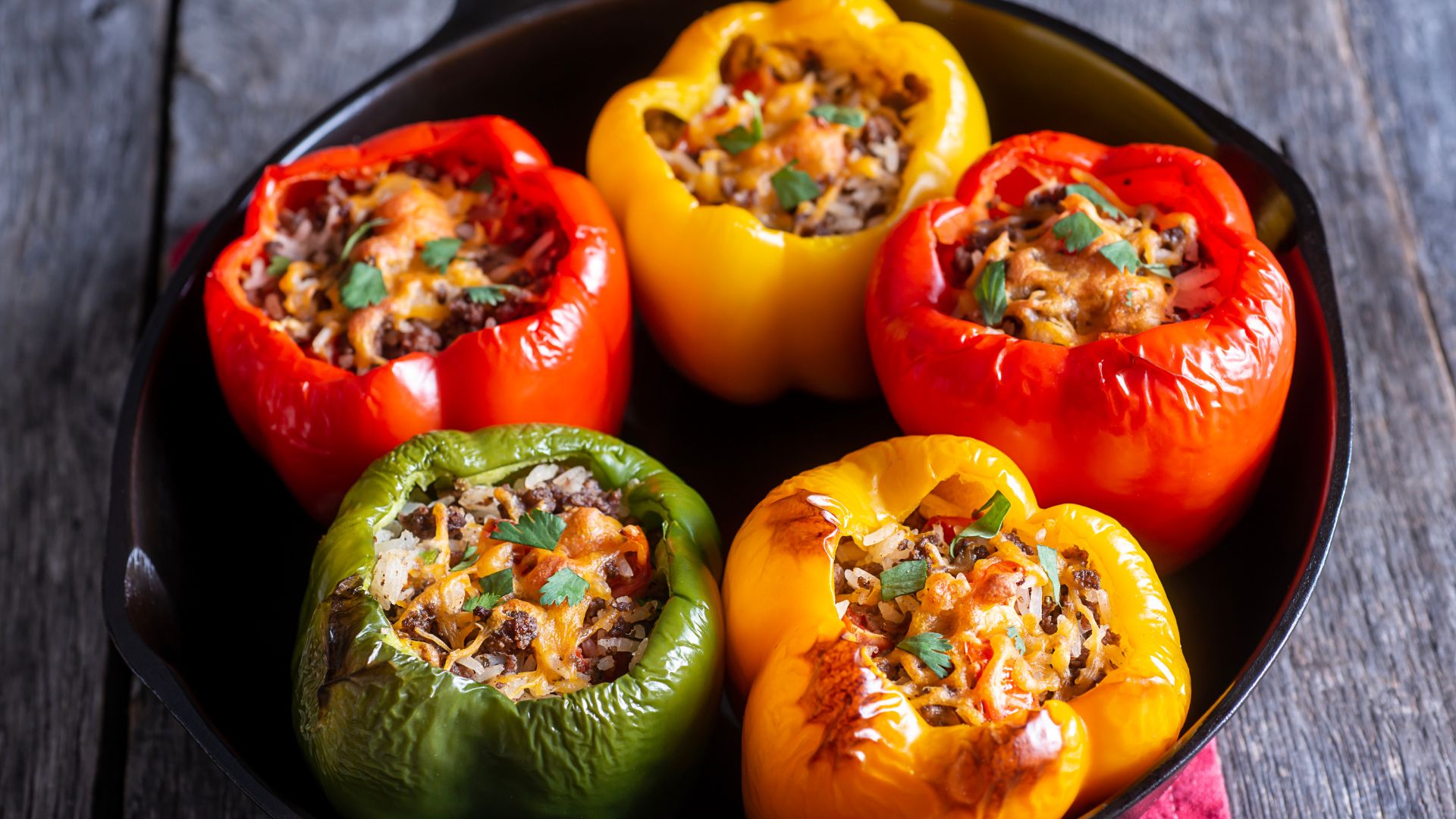 A close-up shot of several vibrant stuffed bell peppers (red, yellow, and green) filled with a savory mixture and topped with melted cheese and herbs, arranged in a dark pan.