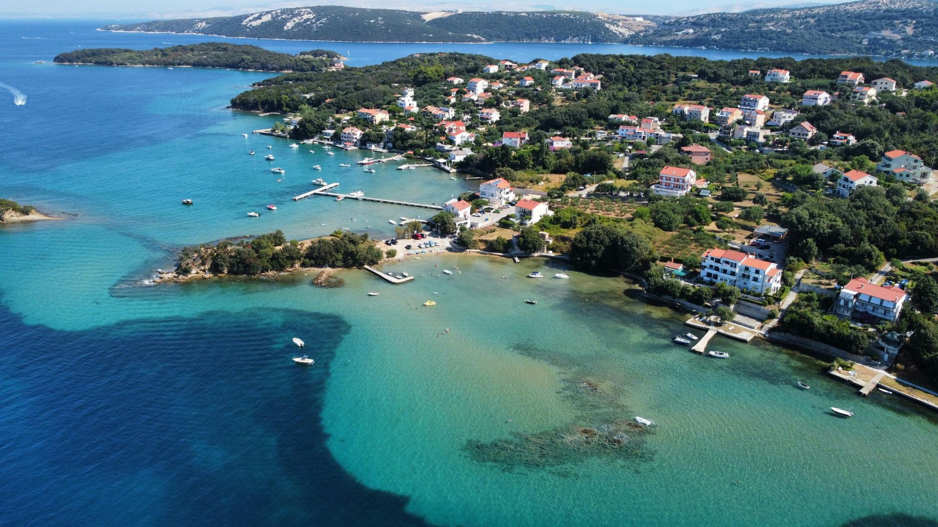 An aerial view of a picturesque coastal village and bay on the island of Rab, Croatia, featuring clear turquoise waters, numerous boats, a small marina, and houses nestled among green hills.