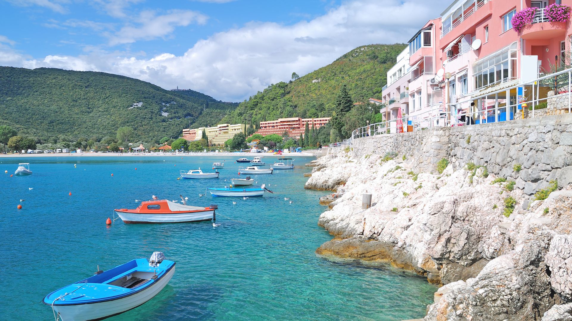 A scenic view of Rabac, Croatia, showing a clear turquoise bay with several small boats anchored in the water, a rocky coastline, and buildings with colorful facades lining the shore against a backdrop of green, forested hills under a blue sky.