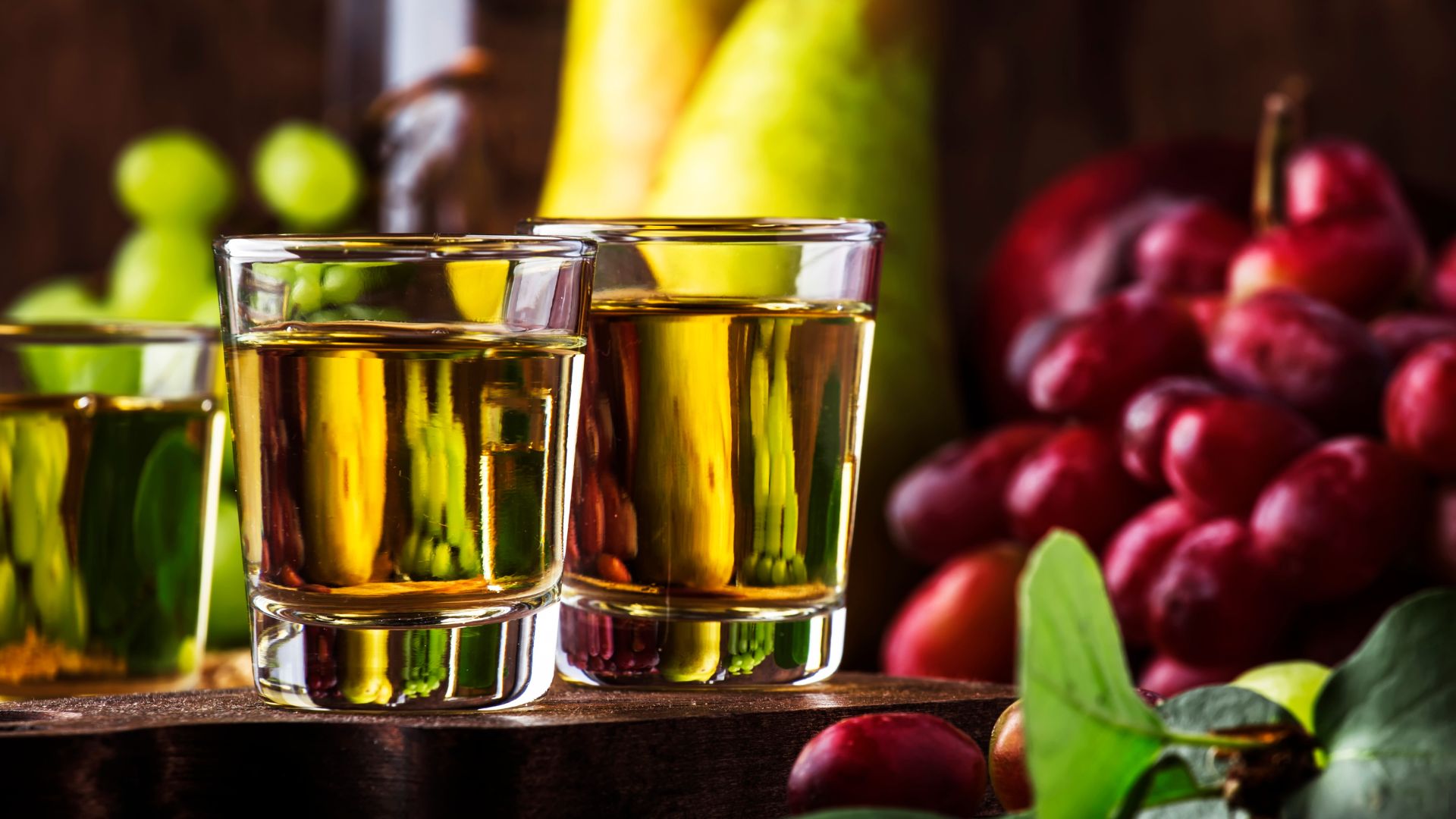 Two glasses of golden-colored spirit, Rakija, on a wooden board with grapes and a pear in the background.