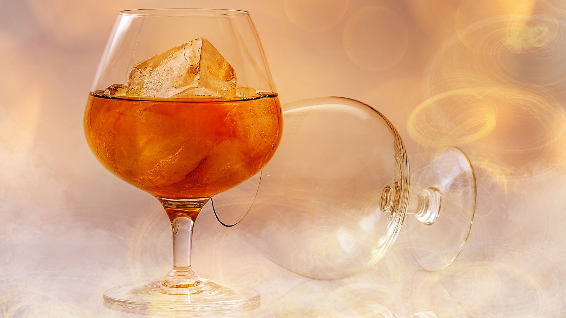 A close-up shot of a snifter glass filled with an amber-colored spirit and ice, with an empty, overturned snifter glass lying beside it on a softly lit, blurred background.