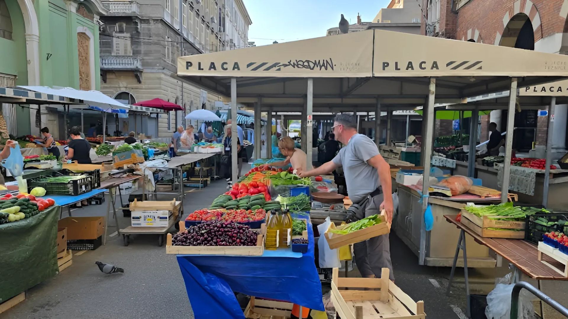 A bustling outdoor market scene in Rijeka, Croatia, with vendors selling fresh produce like fruits and vegetables under canopied stalls, and customers browsing the offerings. Old buildings line the street behind the market, and a pigeon walks on the ground in the foreground.