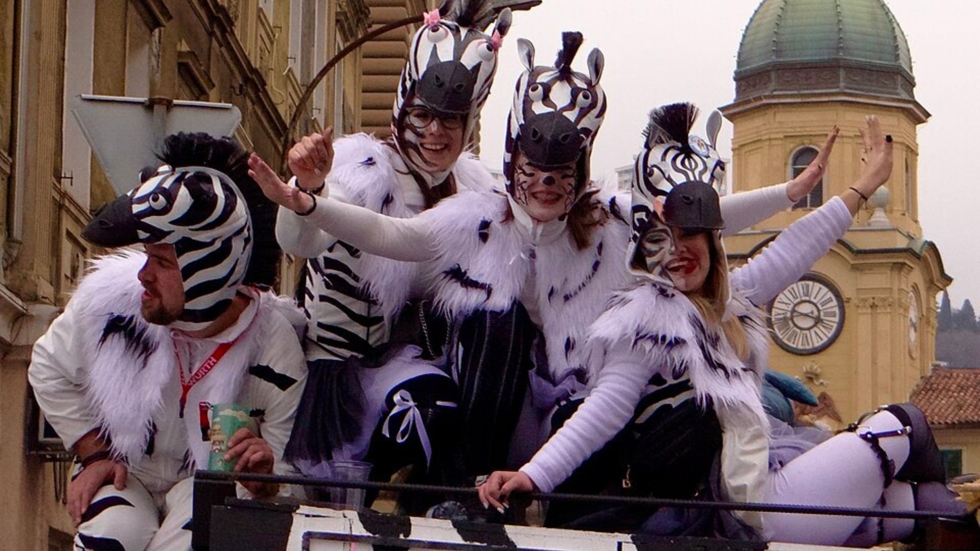 A group of five people in zebra-themed costumes, including striped masks and white furry outfits, are smiling and gesturing on what appears to be a parade float or vehicle, with a building and a clock tower in the background under an overcast sky.