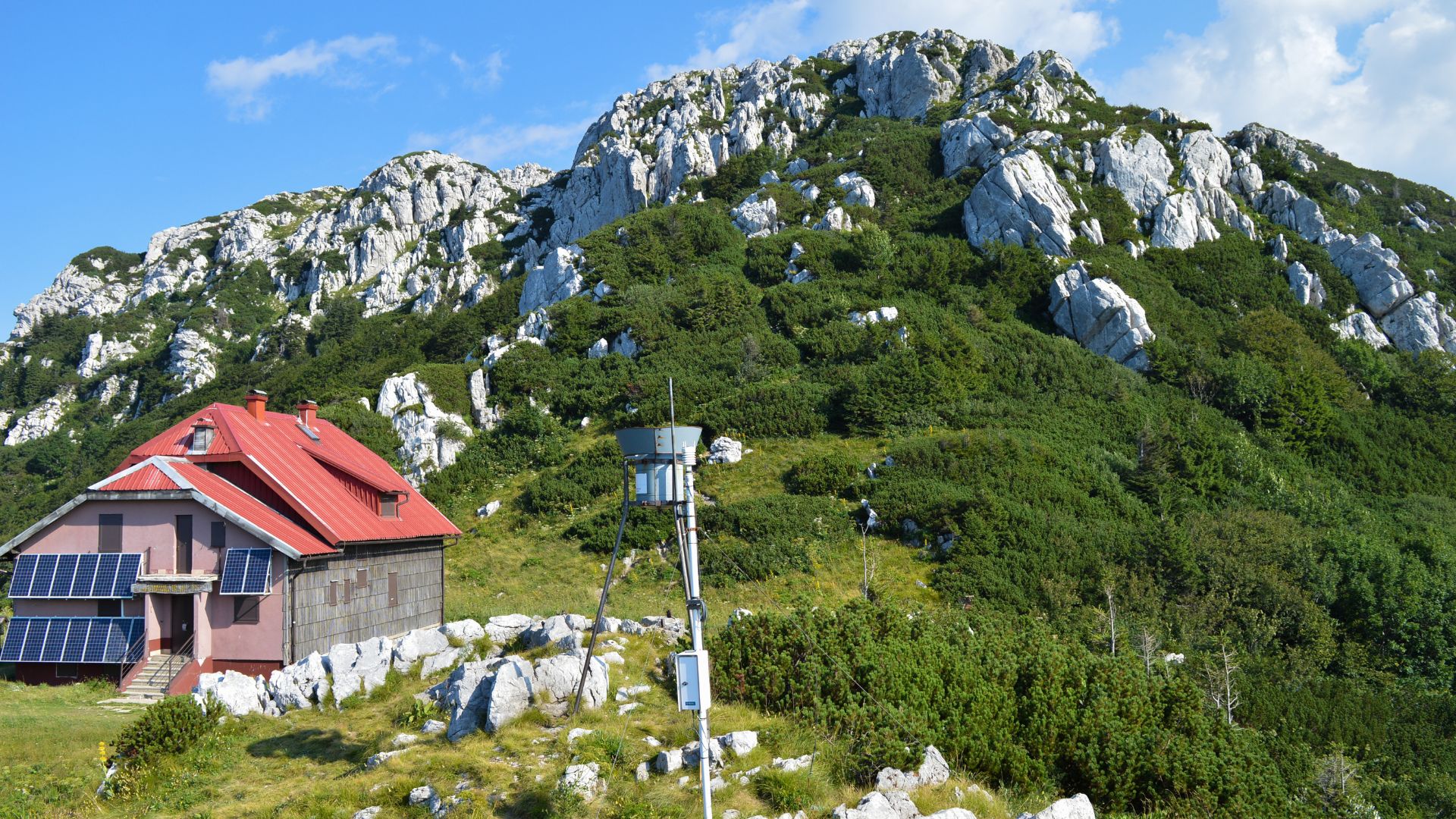 A mountain hut with a red roof and solar panels on its side sits on a rocky, grassy slope, with a weather station nearby. Behind it, a large, rugged mountain covered in dense green vegetation and rocky outcrops rises under a clear blue sky.