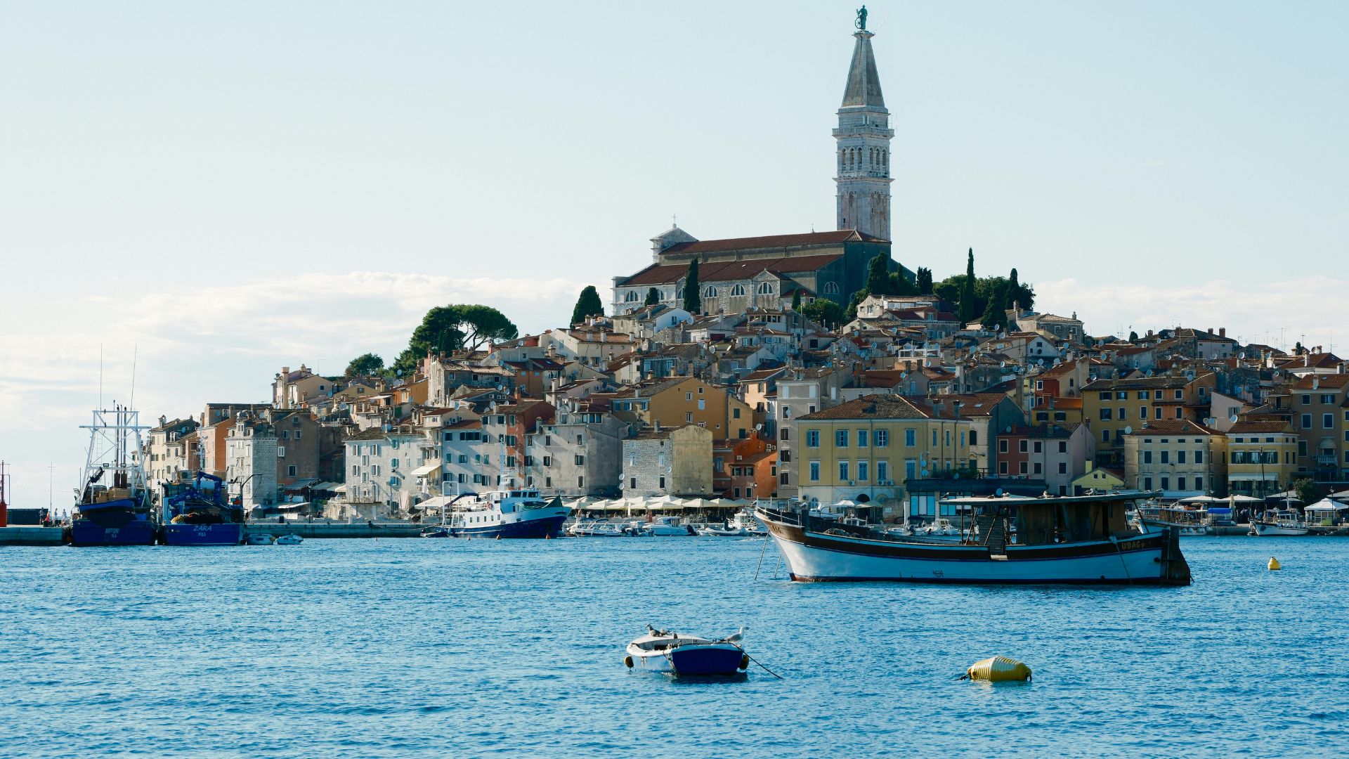 A picturesque view of the coastal town of Rovinj, Croatia, with colorful buildings and a prominent church tower on a hill overlooking the clear blue Adriatic Sea, dotted with boats in the foreground.