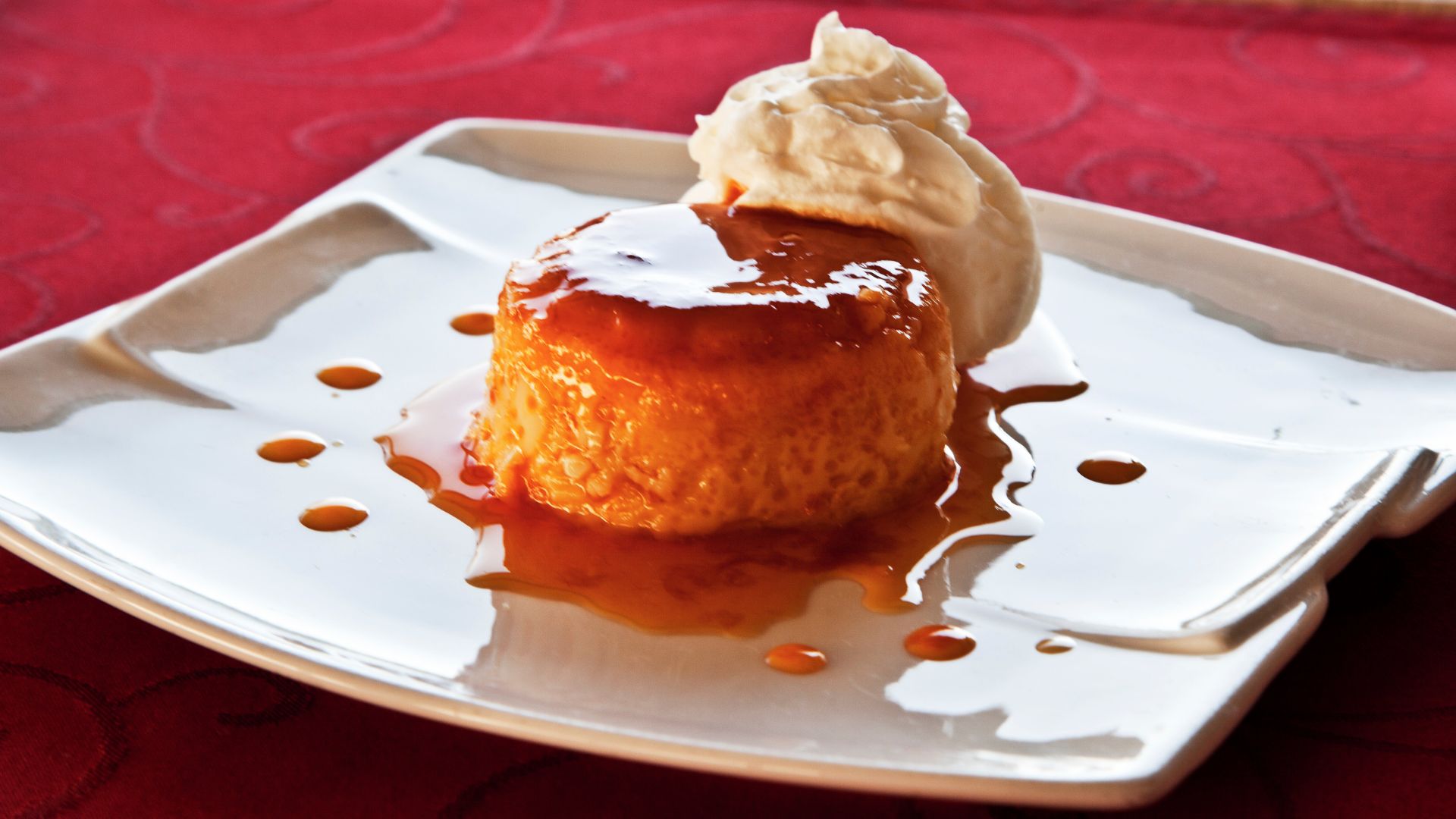 A close-up shot of a golden-brown crème caramel or flan, glistening with caramel sauce, served on a white plate and topped with a dollop of whipped cream.