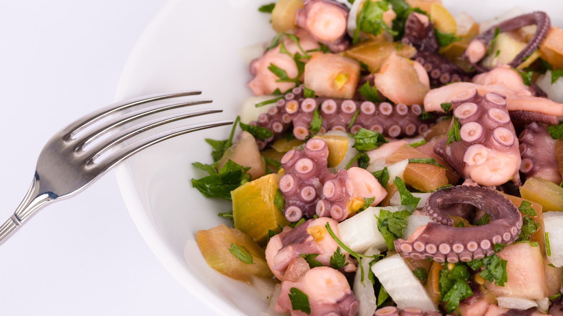 A close-up of a white bowl filled with octopus salad, featuring cooked octopus pieces, diced potatoes, and chopped green herbs like parsley, with a fork resting on the edge of the bowl. 