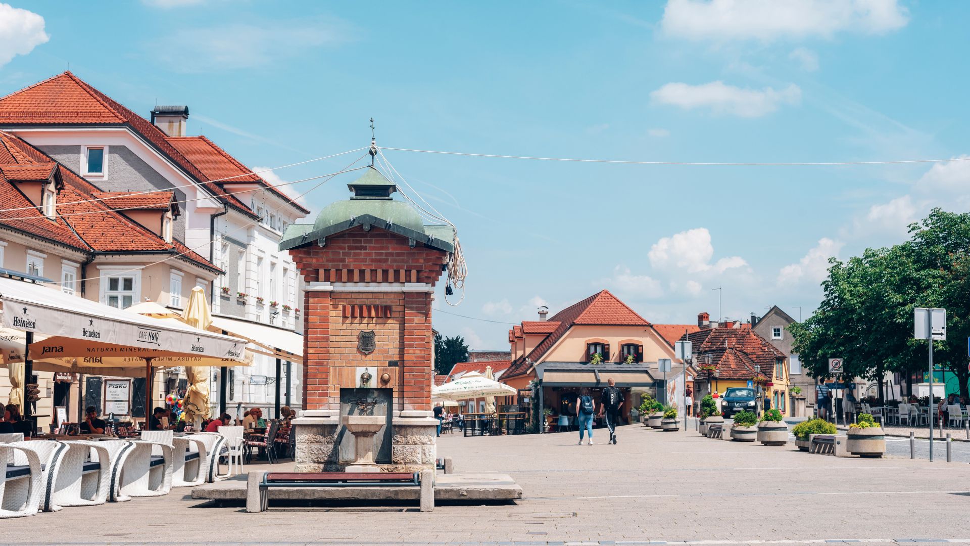A sunny daytime view of a bustling European town square featuring a prominent brick fountain in the center, surrounded by historic buildings with red-tiled roofs, outdoor cafes with umbrellas, and a few pedestrians walking in the distance.