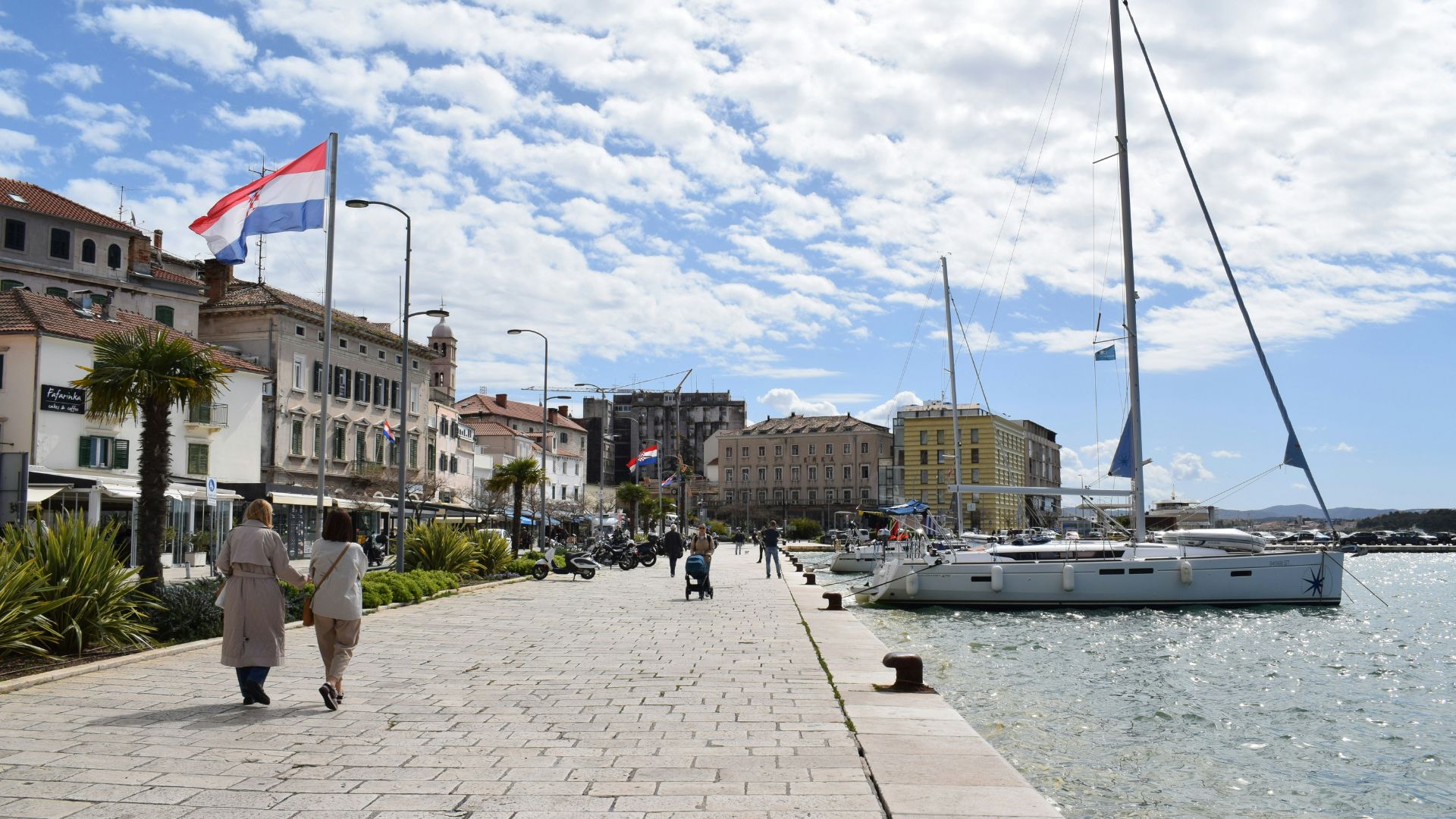 A sunny day view of the Šibenik promenade in Croatia, with a wide, paved walkway alongside a calm body of water featuring several sailboats docked at the pier. On the left, historic buildings with colorful facades line the promenade, and two individuals are seen walking away from the viewer. A Croatian flag flies prominently above the buildings on the left.