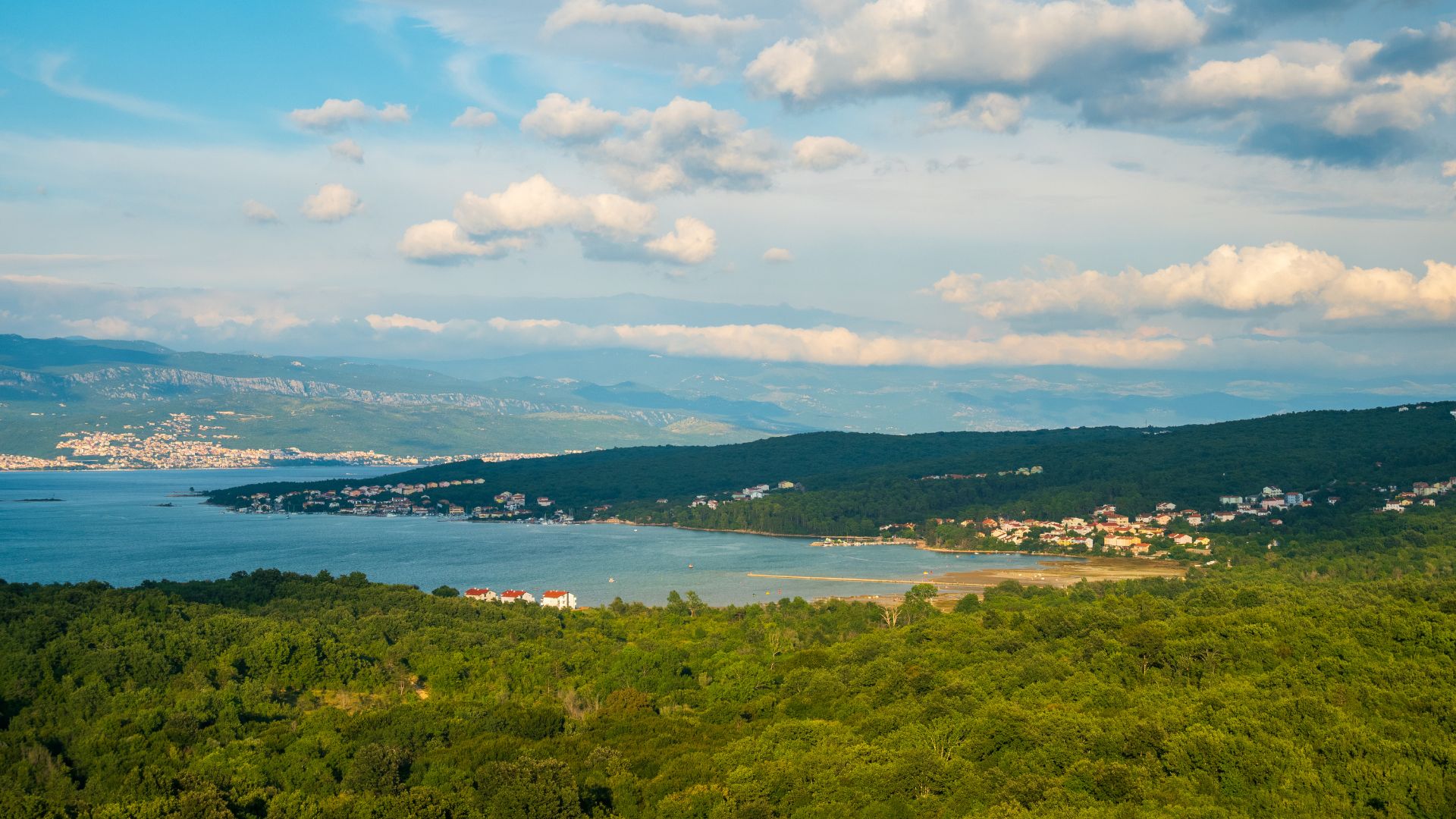 A panoramic view of a serene bay with turquoise water, surrounded by dense green forests and hills, with scattered houses and a small town visible along the coastline under a partly cloudy sky.