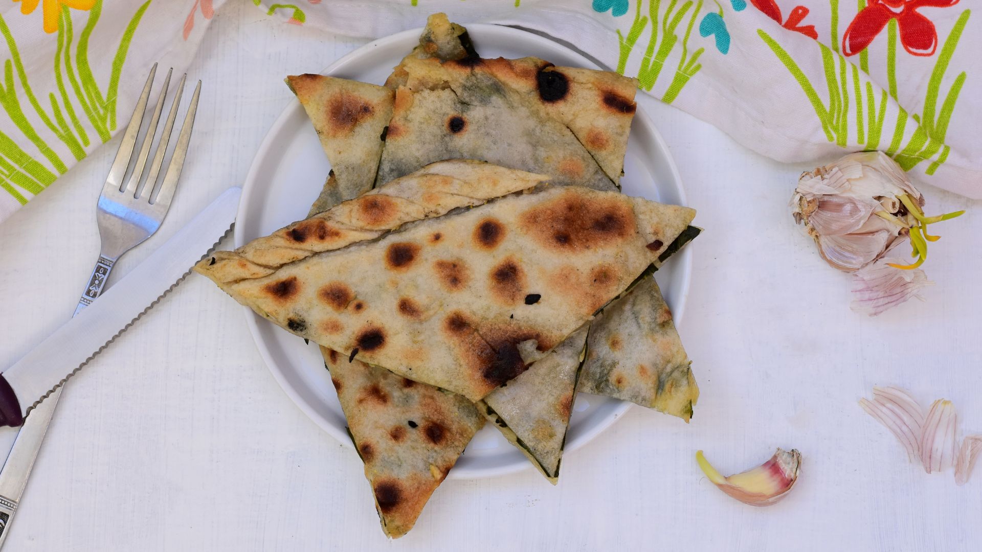 A plate of baked Soparnik, a traditional Dalmatian flatbread filled with greens, served alongside garlic cloves on a white surface with a patterned cloth in the background.
