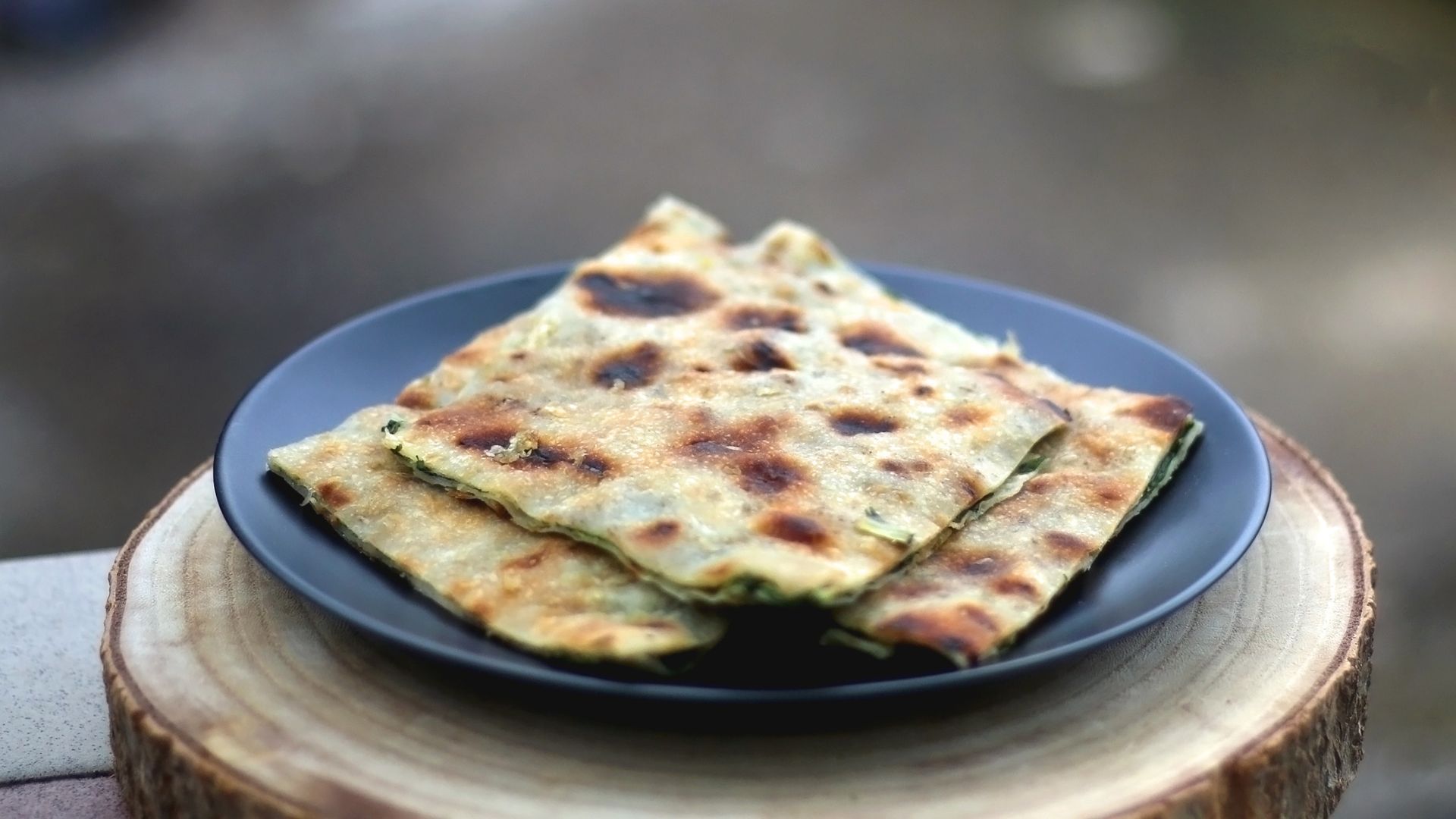 A close-up shot of several pieces of golden-brown Soparnik, a traditional Croatian savory pie filled with Swiss chard, garlic, and olive oil, arranged on a dark plate, resting on a wooden tree stump slice. 