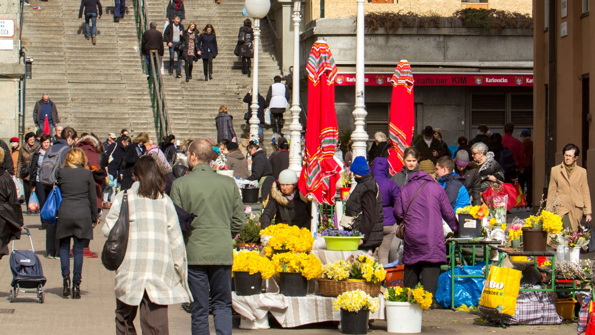 A busy outdoor flower market scene with vendors selling yellow flowers in buckets and baskets, surrounded by numerous people walking and shopping, with stairs and buildings visible in the background.