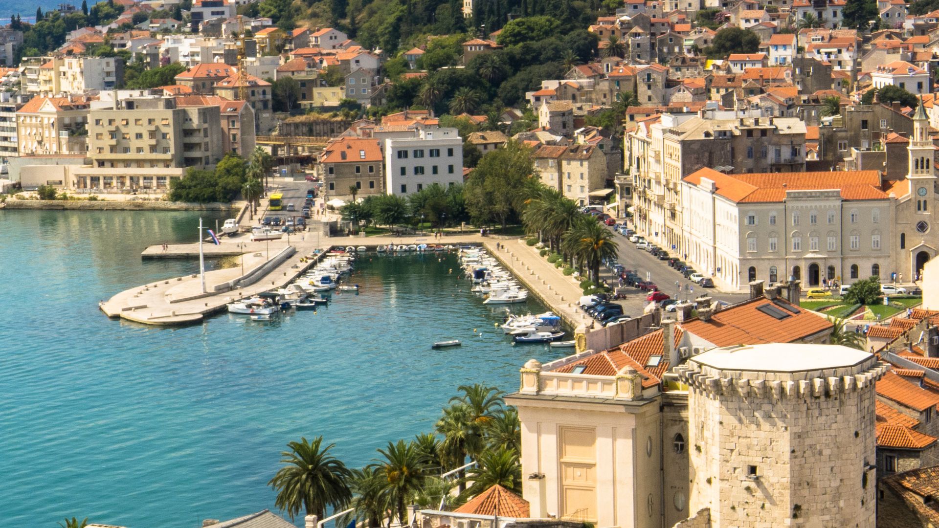 An aerial view of a vibrant coastal city in Europe with a bustling harbor filled with boats, ancient buildings, and a prominent historical palace complex, likely Diocletian's Palace in Split, Croatia, set against a backdrop of hills and blue water.