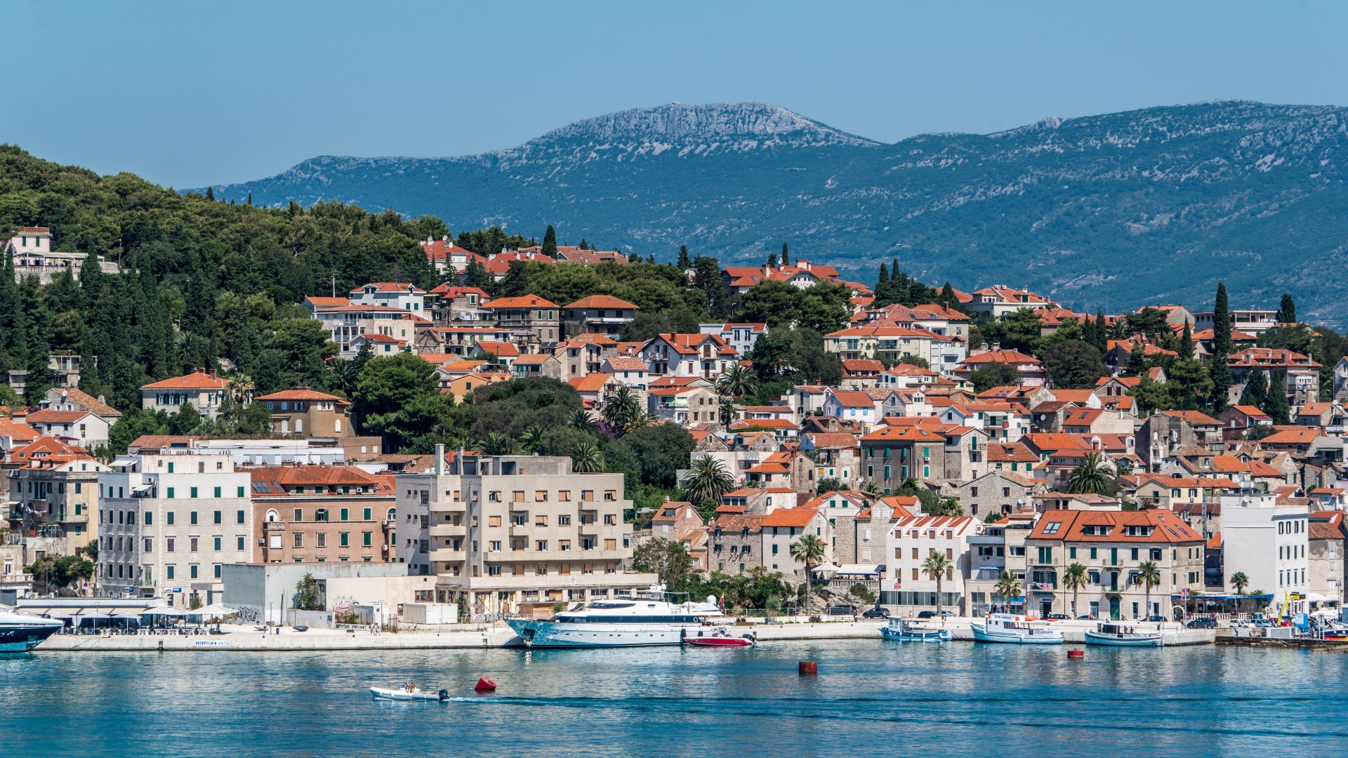 A panoramic view of Split, Croatia, showing a bustling waterfront with boats docked in the clear blue Adriatic Sea, framed by a hillside covered with traditional red-roofed buildings and a distant mountain range under a clear sky.