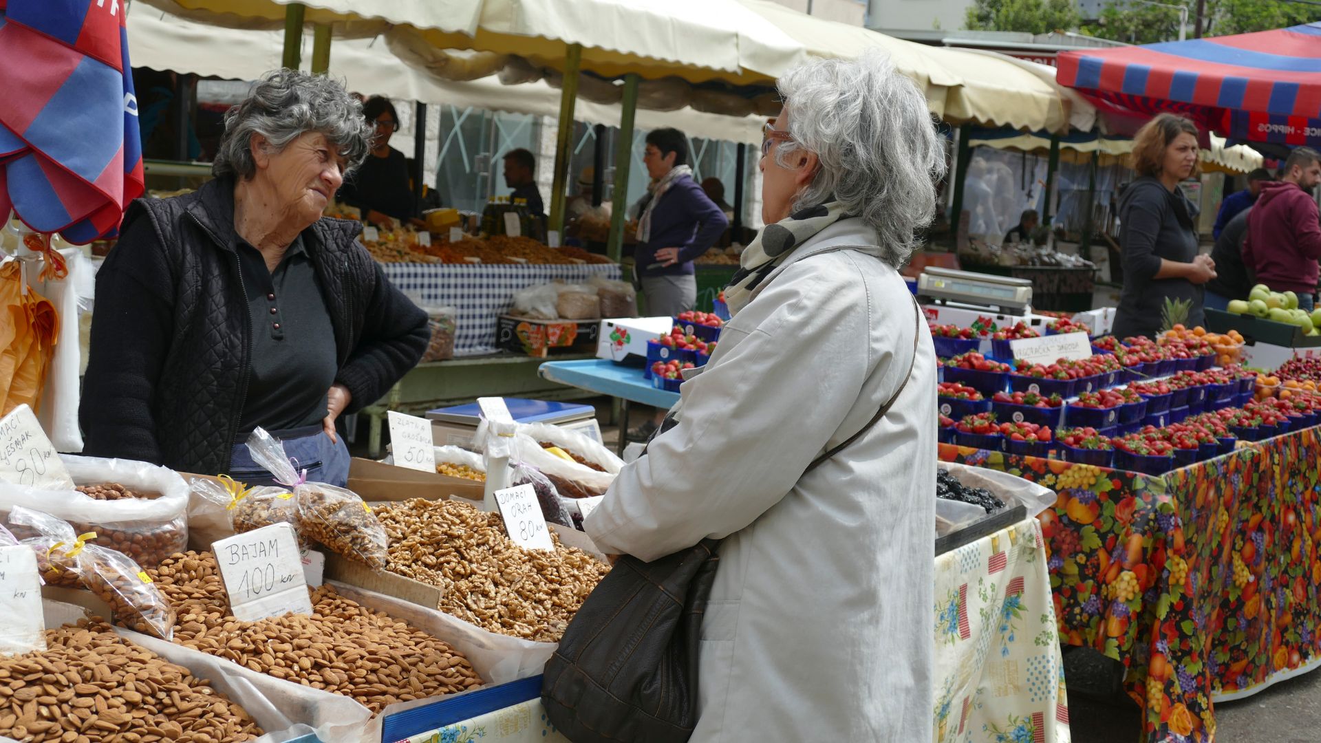 A vibrant outdoor market scene where two women are conversing at a stall laden with nuts and dried fruits, while other market stalls with colorful umbrellas and fresh produce are visible in the background.