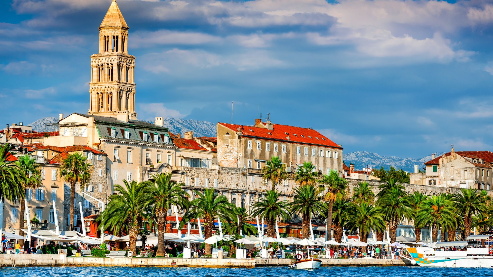 A panoramic view of the historic city of Split, Croatia, showing the bell tower of the Cathedral of Saint Domnius and the ancient architecture of Diocletian's Palace along the waterfront, with palm trees in the foreground and mountains in the background under a partly cloudy sky.