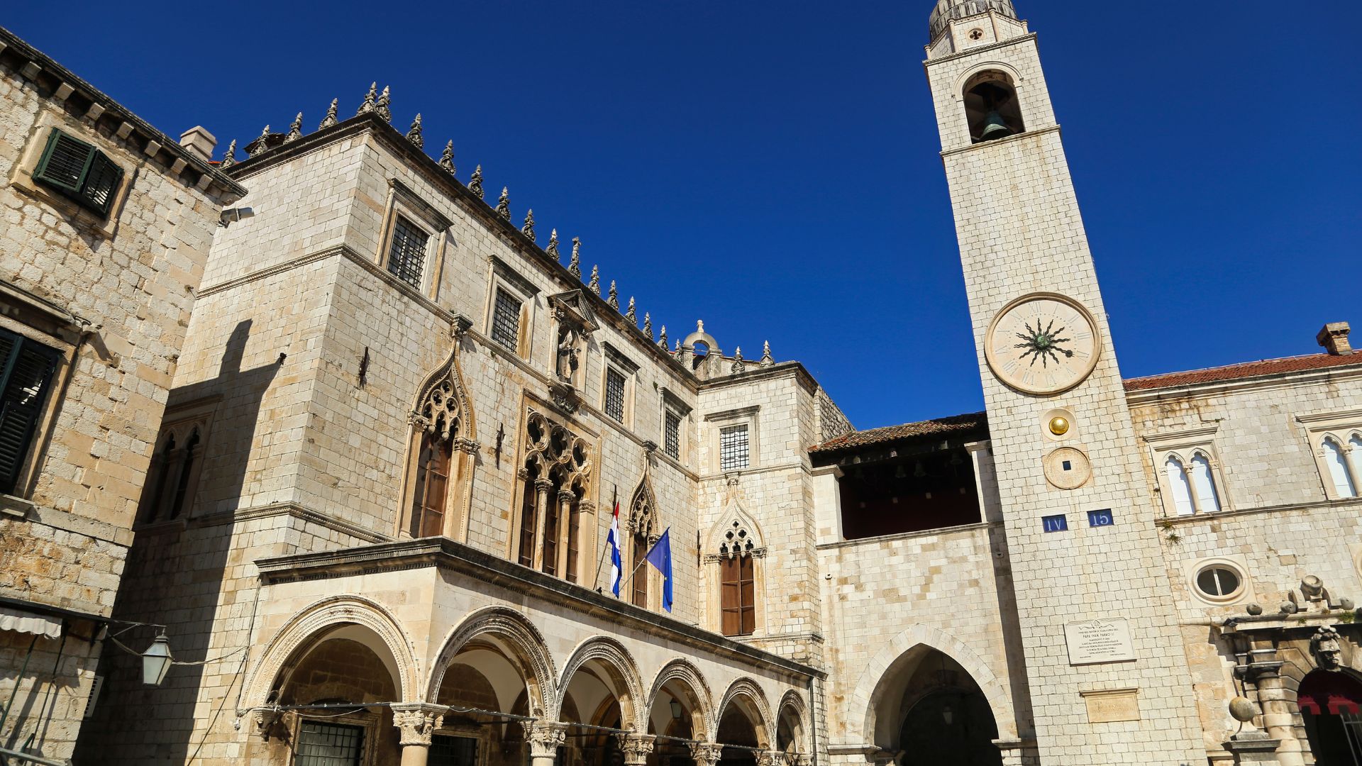 A low-angle shot on a bright, clear day of the Sponza Palace and the Bell Tower in Dubrovnik, Croatia, showcasing their distinct Gothic and Renaissance architectural elements and the prominent clock face on the tower.